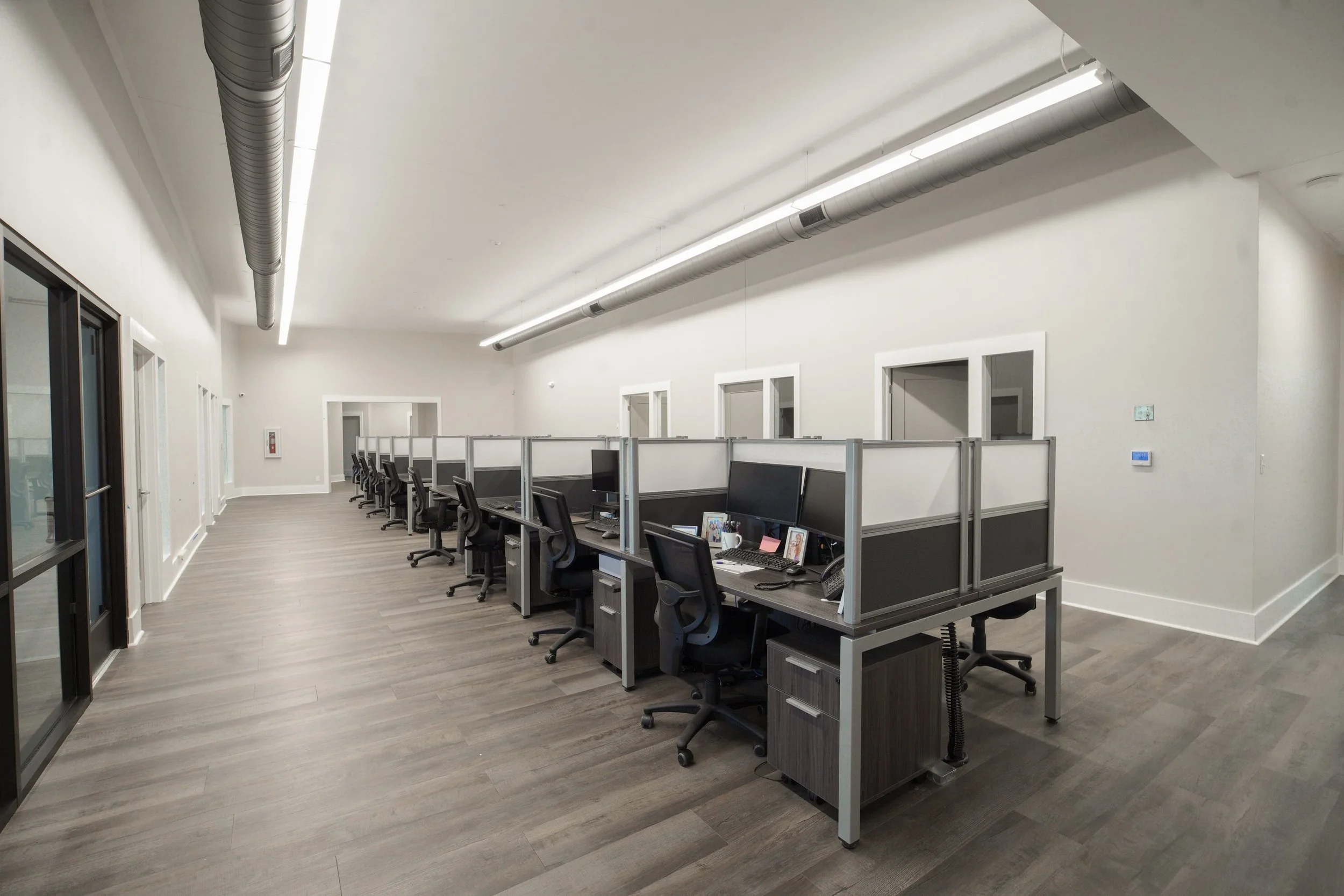 An office with a row of workstations and chairs, gray wooden floors, white walls, modern lighting, and wall-mounted air ducts.