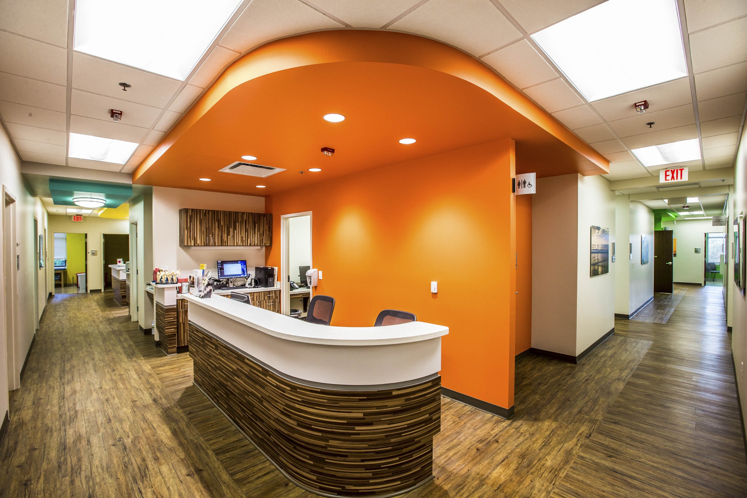 Hospital reception area with a curved front desk, orange accent wall, and wood flooring, featuring ceiling lights and signs for restrooms and exits.