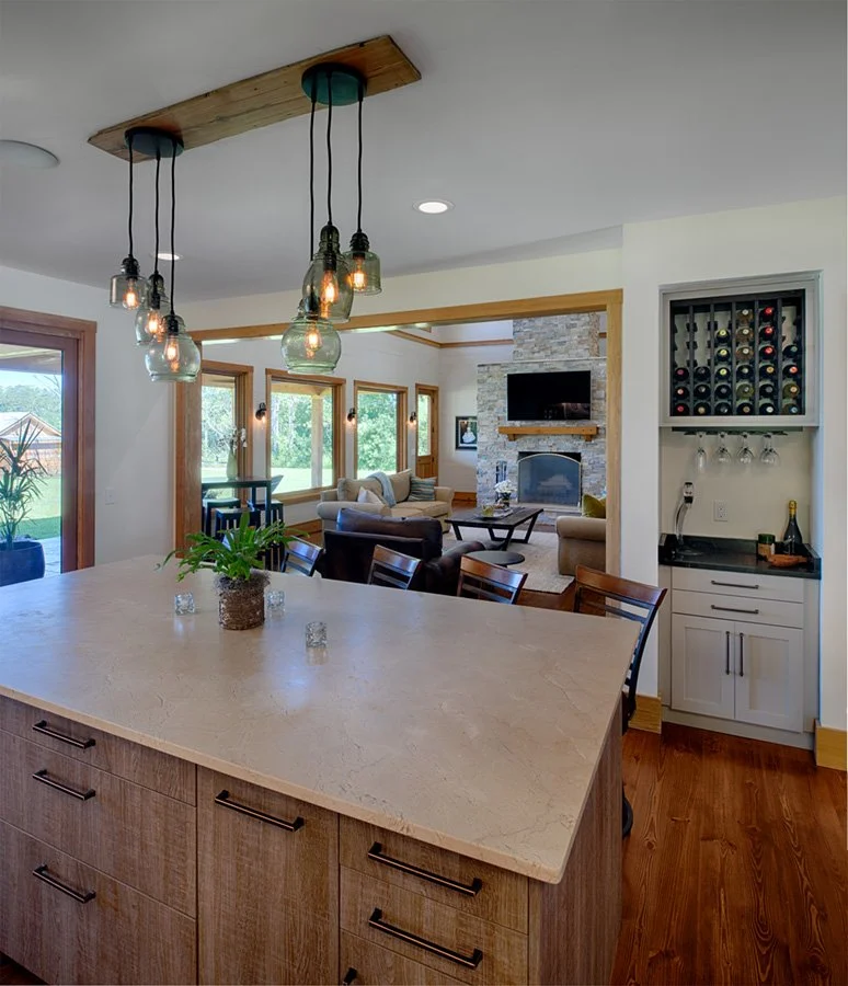Open-concept living space with a kitchen island featuring a beige countertop, hanging pendant lights, a dining area with wooden chairs, a cozy living room with multiple sofas, a stone fireplace, a wall-mounted TV, and large windows letting in natural