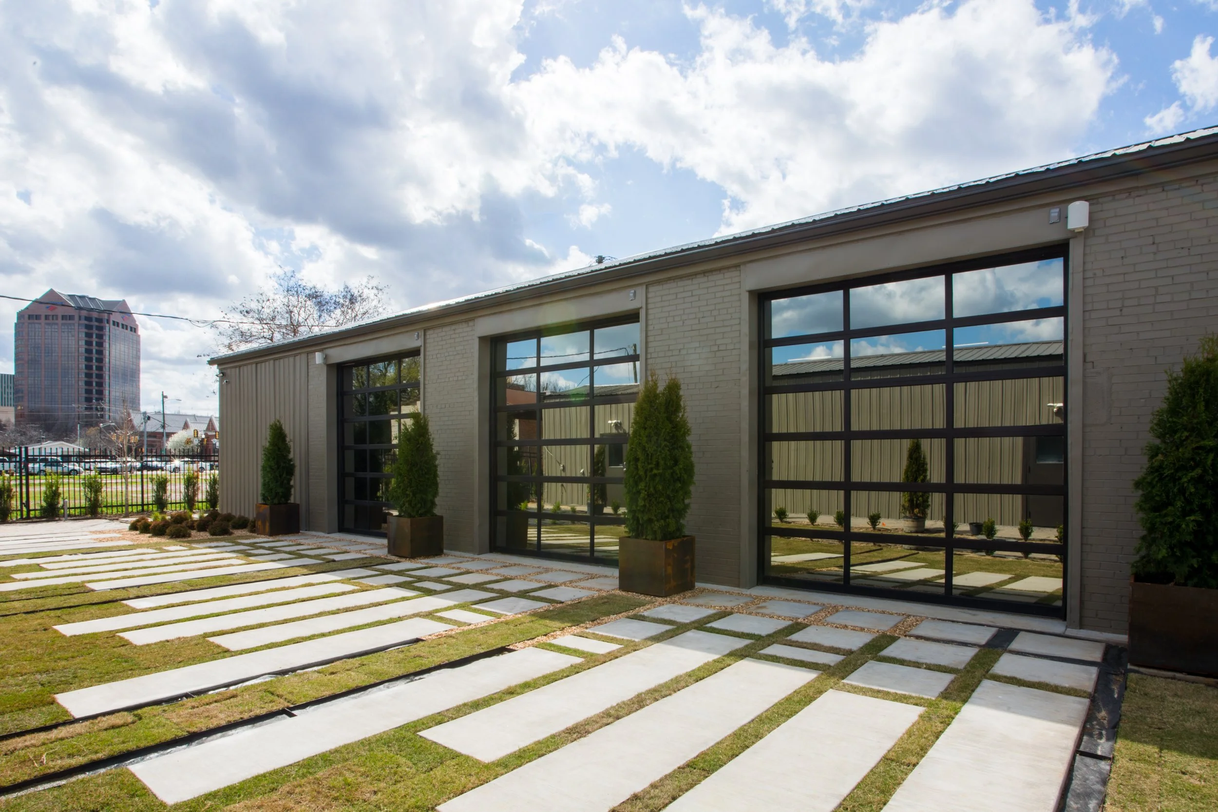 Modern building with large glass windows and a decorative walkway with white concrete slabs separated by grass, surrounded by potted plants, under a partly cloudy sky with a cityscape in the background.