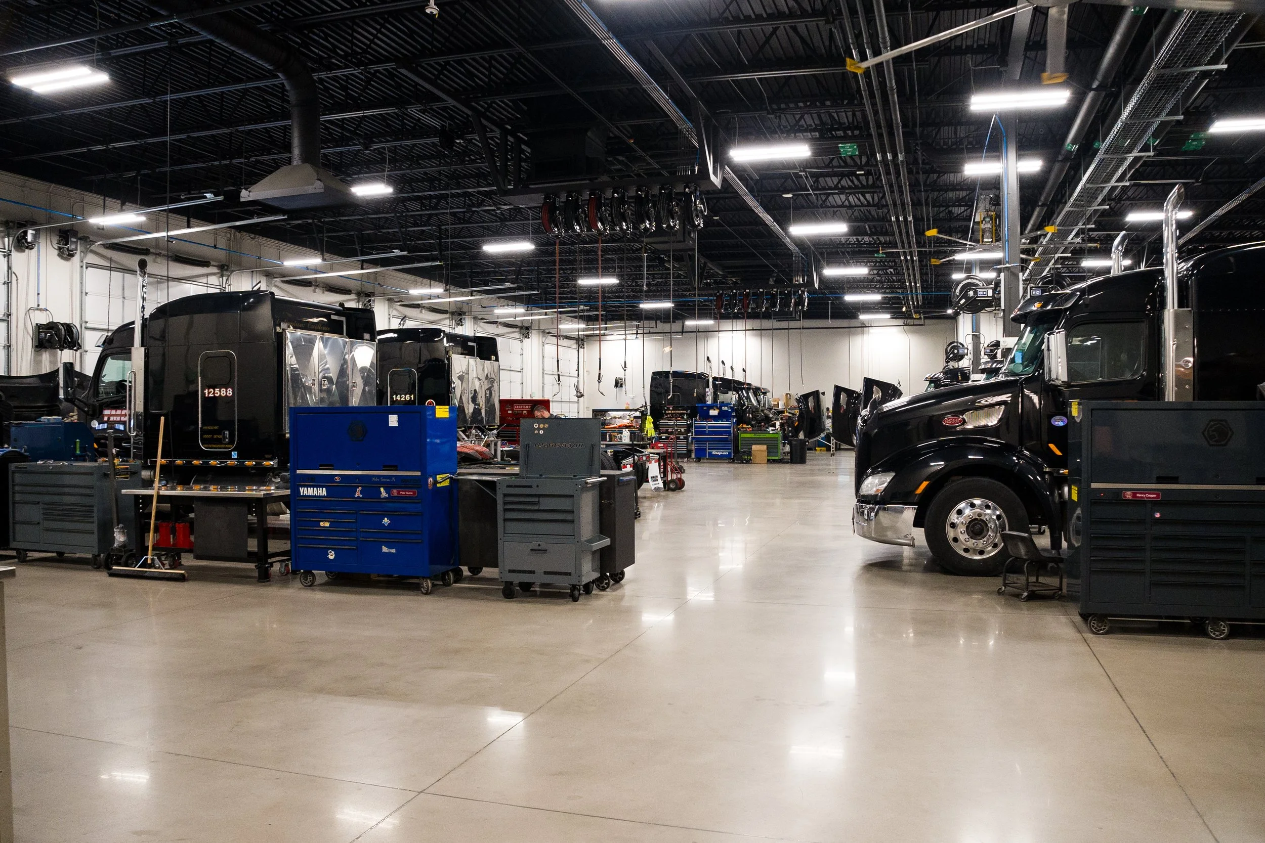 An indoor vehicle service garage with black semi-trucks parked inside, various tools, toolboxes, and equipment are scattered around, and the ceiling has industrial lighting and exposed ductwork and piping.
