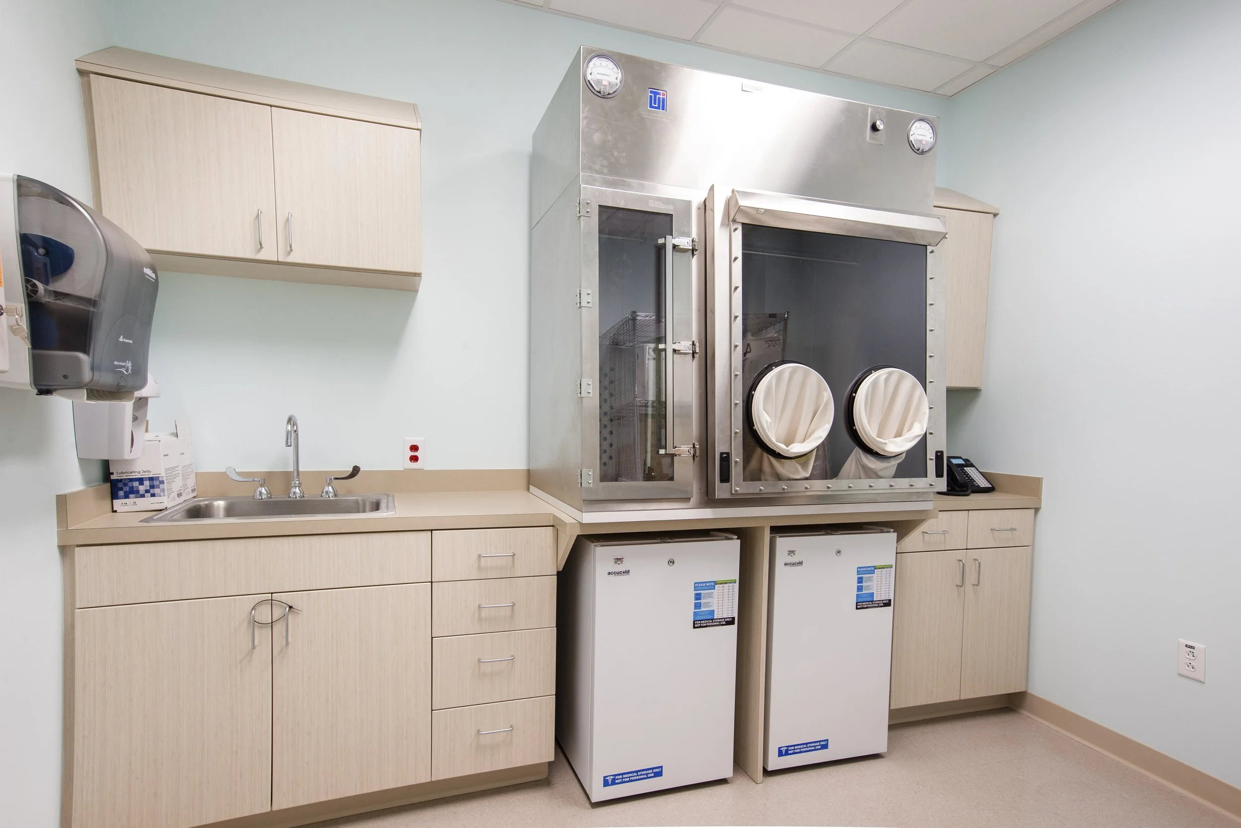 Medical sterilization room with a stainless steel sterilizer, a nearby sink, white cabinets above and below, and disinfectant bottles on the counter.