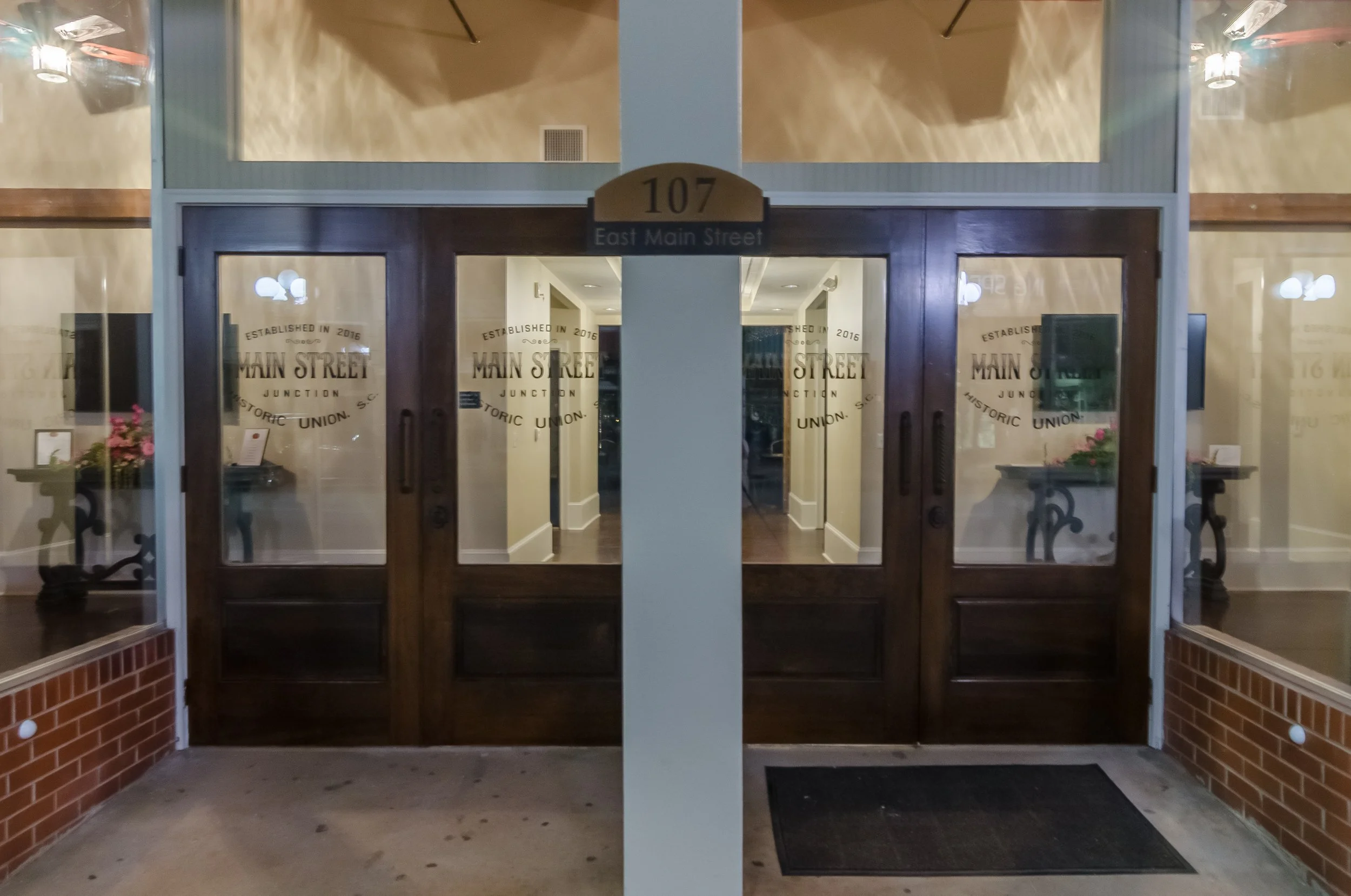 Double glass door entrance with wooden frames, above which there is a sign with the number 107 and the words 'East Main Street'. Inside, the hallway is well-lit with ceiling lights, and there are glass display cases on either side with pink flowers i