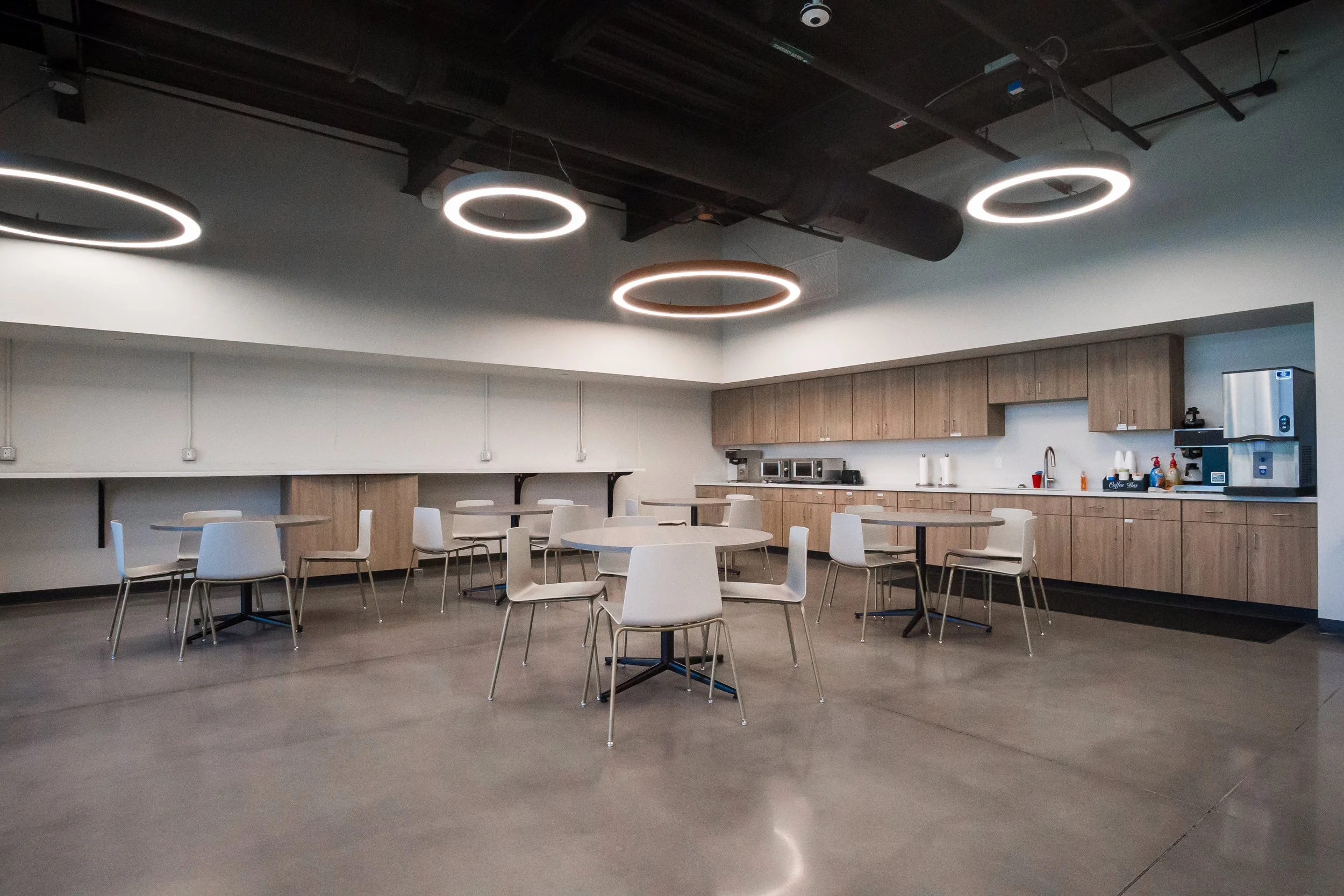 Modern cafeteria with white round tables and white chairs, wooden cabinets along the back wall, and circular LED ceiling lights.