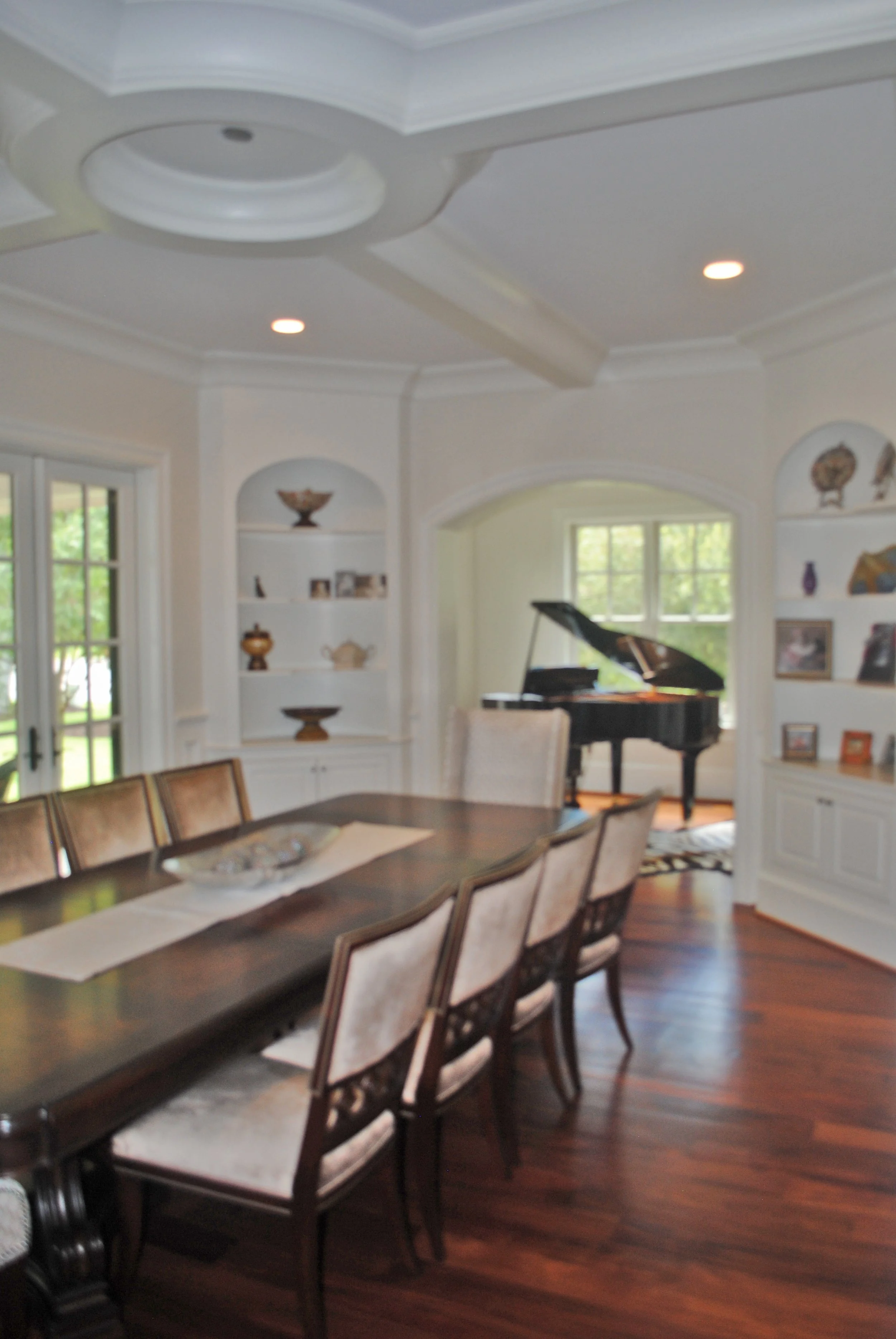 A dining room with a long dark wood table surrounded by chairs. In the background, there is a black grand piano and large windows with green trees outside. The room has built-in white shelves with decorative items and a ceiling with recessed lighting