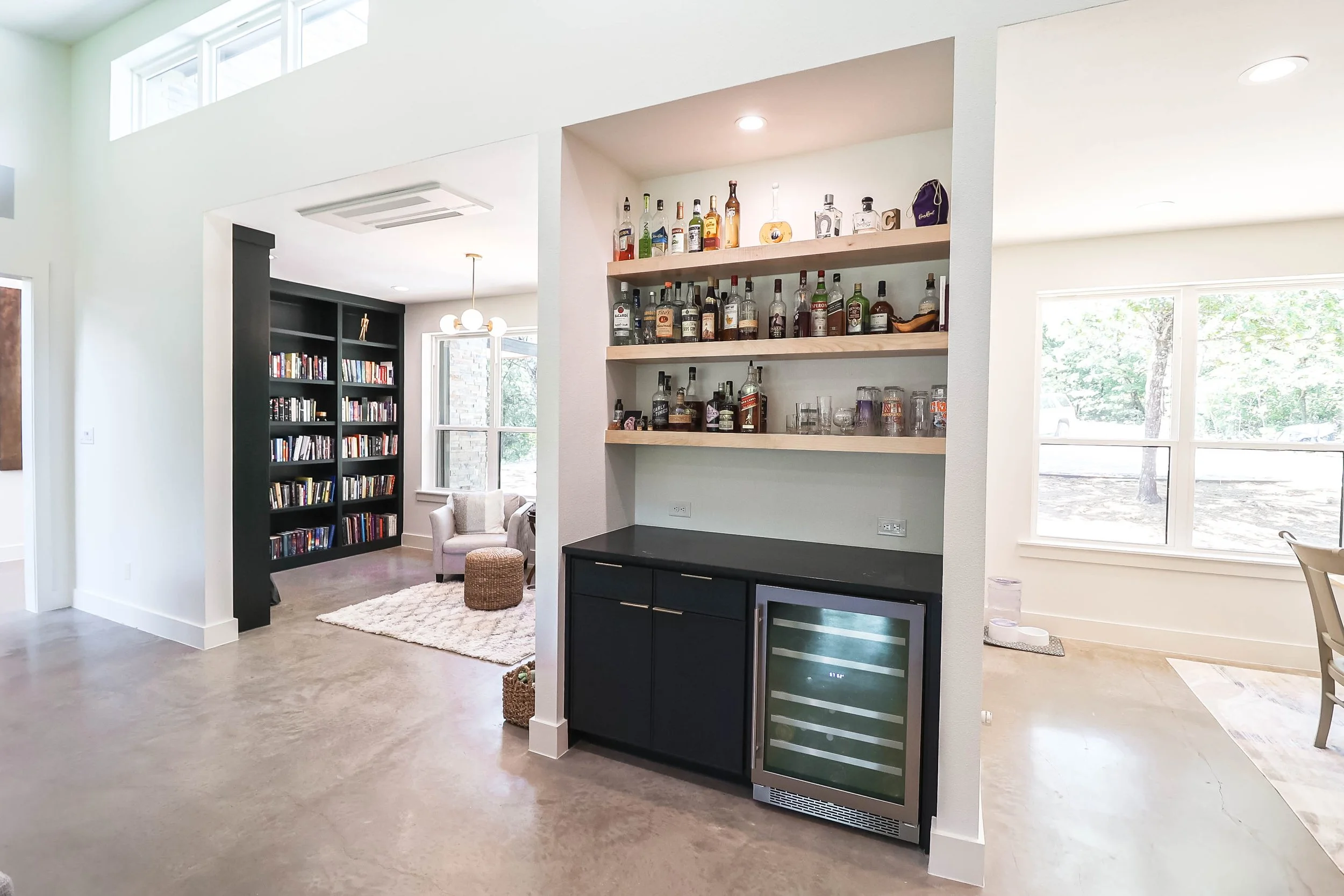 Open living area with bar shelves filled with liquor bottles, a black wine cooler, and a view of a sitting nook with a plush chair, small ottoman, and carpet. Large windows let in natural light.