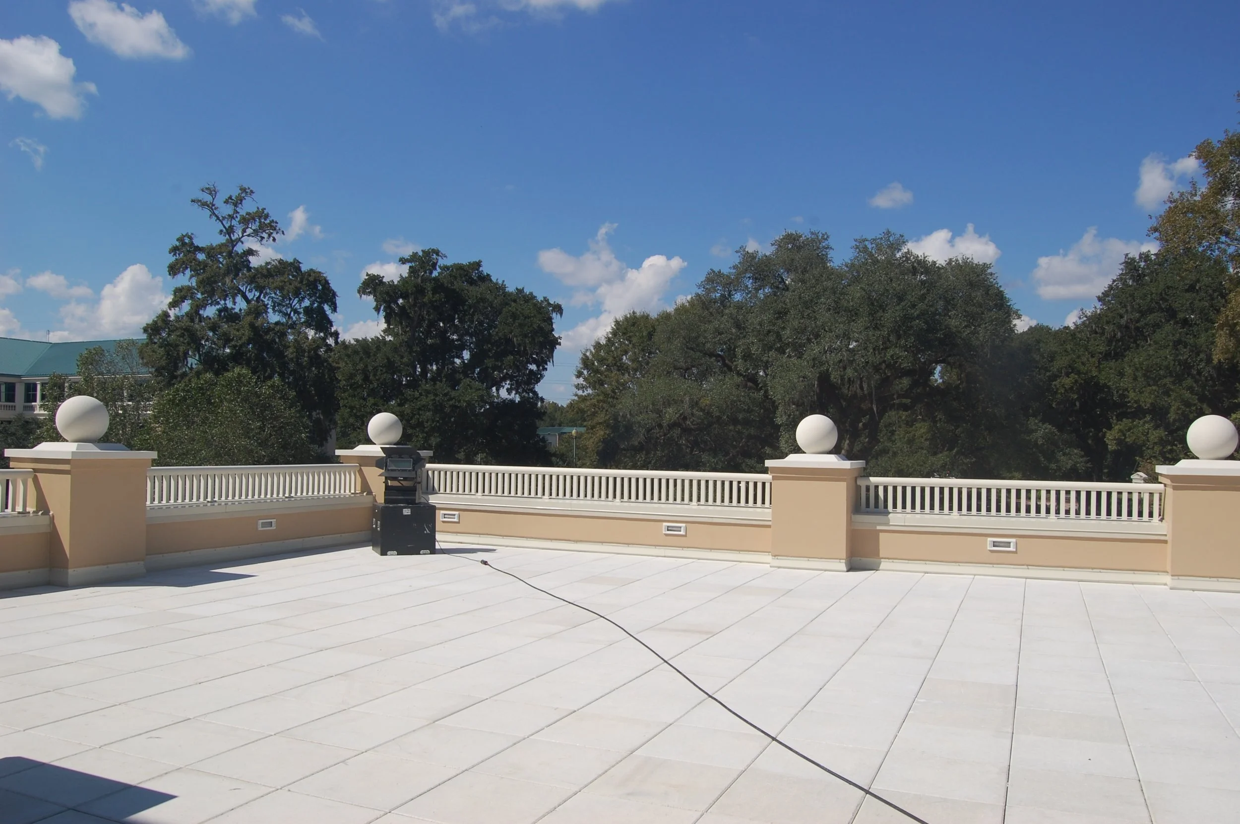 An outdoor terrace with white tiled flooring, decorative white railing with round finials, and a large tree with lush green foliage in the background; blue sky with scattered clouds overhead.