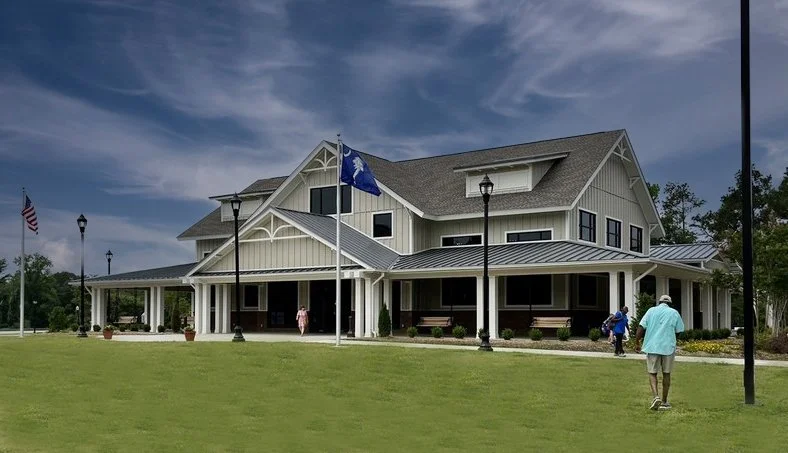 A large, two-story house with a metal roof and white siding, surrounded by a well-maintained green lawn and several lampposts. Three people are walking near the house, and it has an American flag and a flag with a blue background and white emblem.