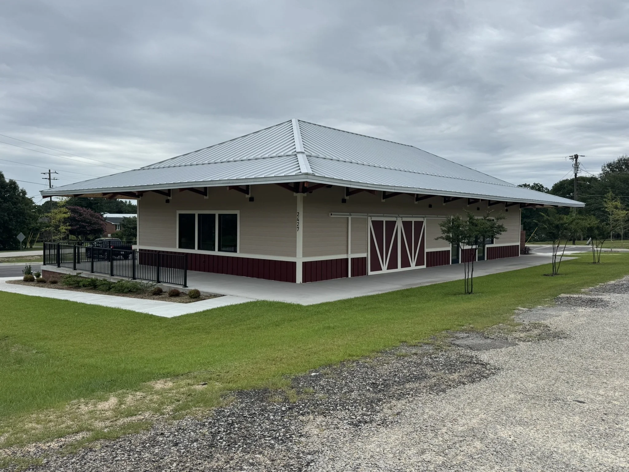 A small building with a metal roof, white walls with beige and burgundy accents, and large barn-style doors. The building is surrounded by a green lawn and trees, with a cloudy sky overhead.