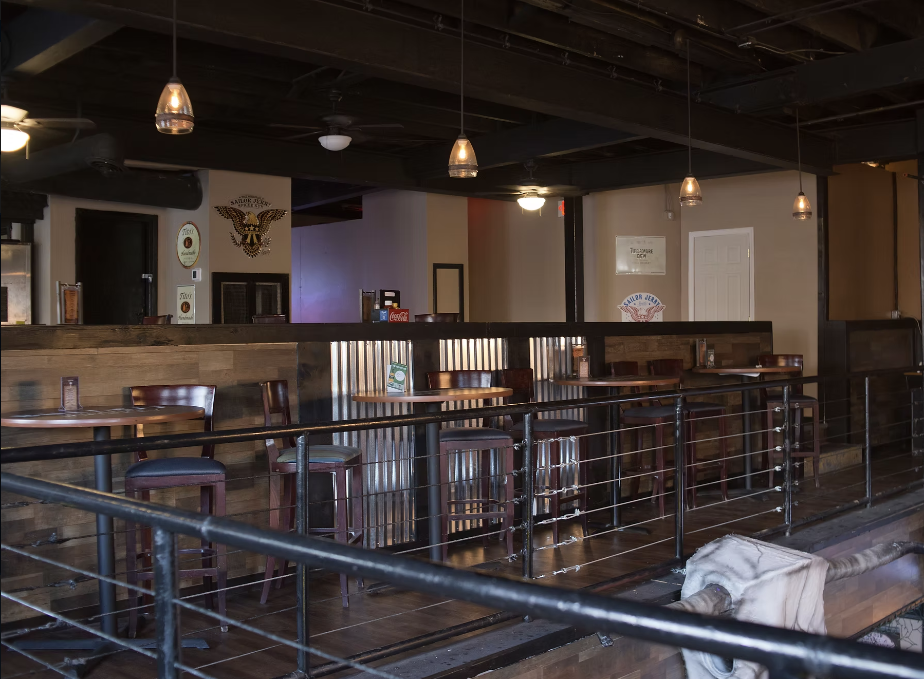 Empty restaurant or bar with wooden tables and chairs, ceiling fans and pendant lights, metal railing, and some signs on the wall.