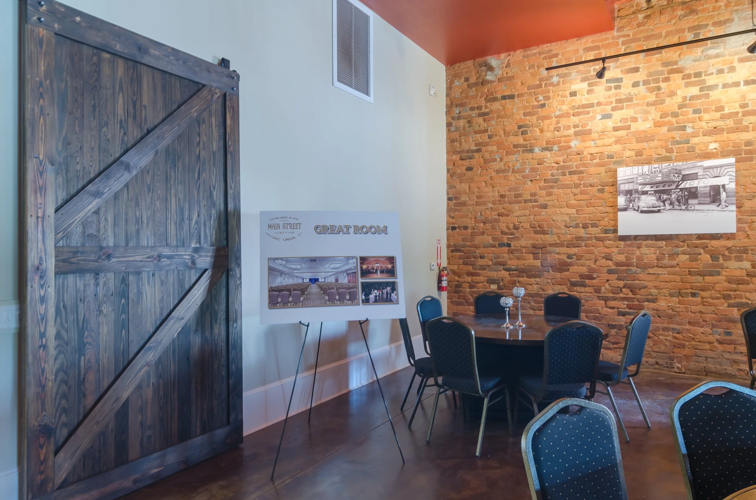 An indoor event space with a brick wall and a white wall, featuring a black and white historical photo, chairs set around a table with decorative glass candle holders, and a large wooden sliding door.