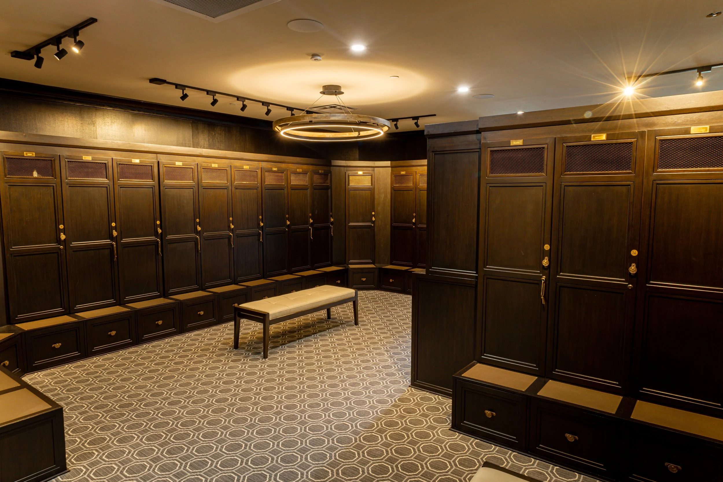 Locker room with dark wooden lockers, a beige bench, and a patterned carpet. Modern ceiling lighting and a circular light fixture are also visible.