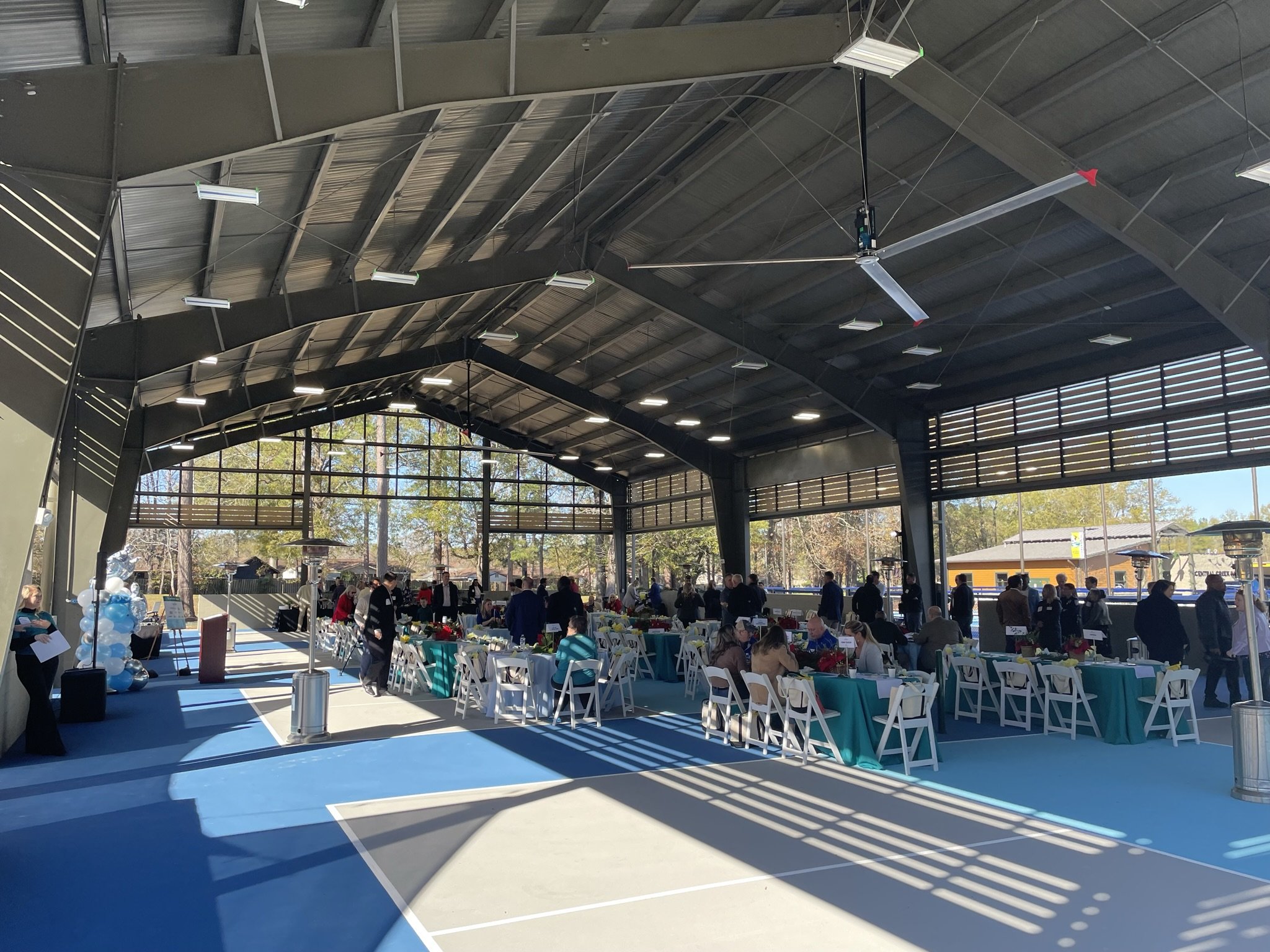 Inside a large, open-air pavilion with a metal roof, decorated tables with teal tablecloths and white chairs are set up for a celebration. People are sitting and standing, engaging in conversations. There are balloons and signs near the entrance, and large windows allow natural light to fill the space.
