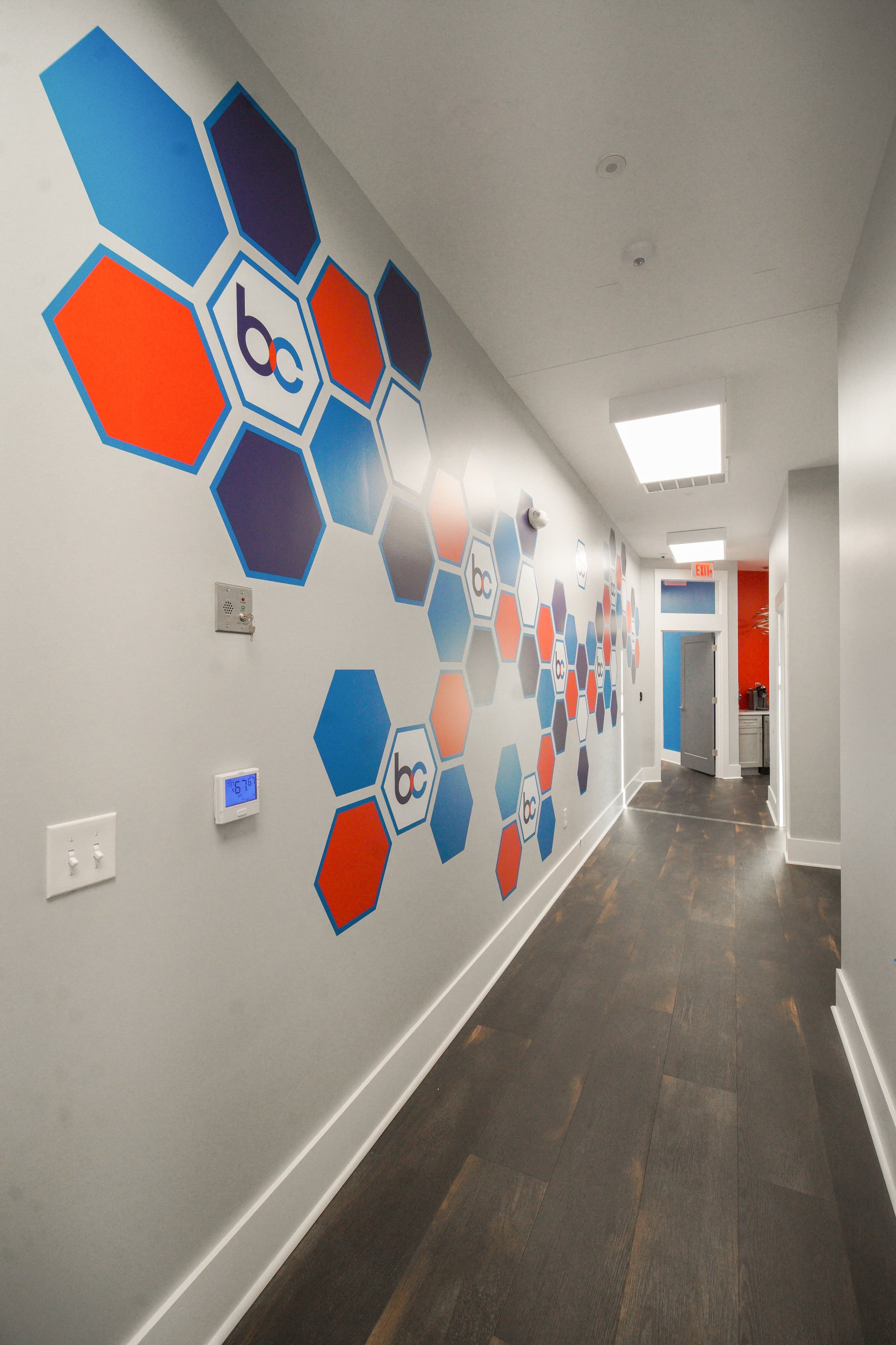 Hallway with white walls and dark hardwood flooring, decorated with a pattern of large hexagonal shapes in blue, orange, and dark purple, containing the letters 'bc' in some of the shapes. The ceiling has bright rectangular lights, and there's a door