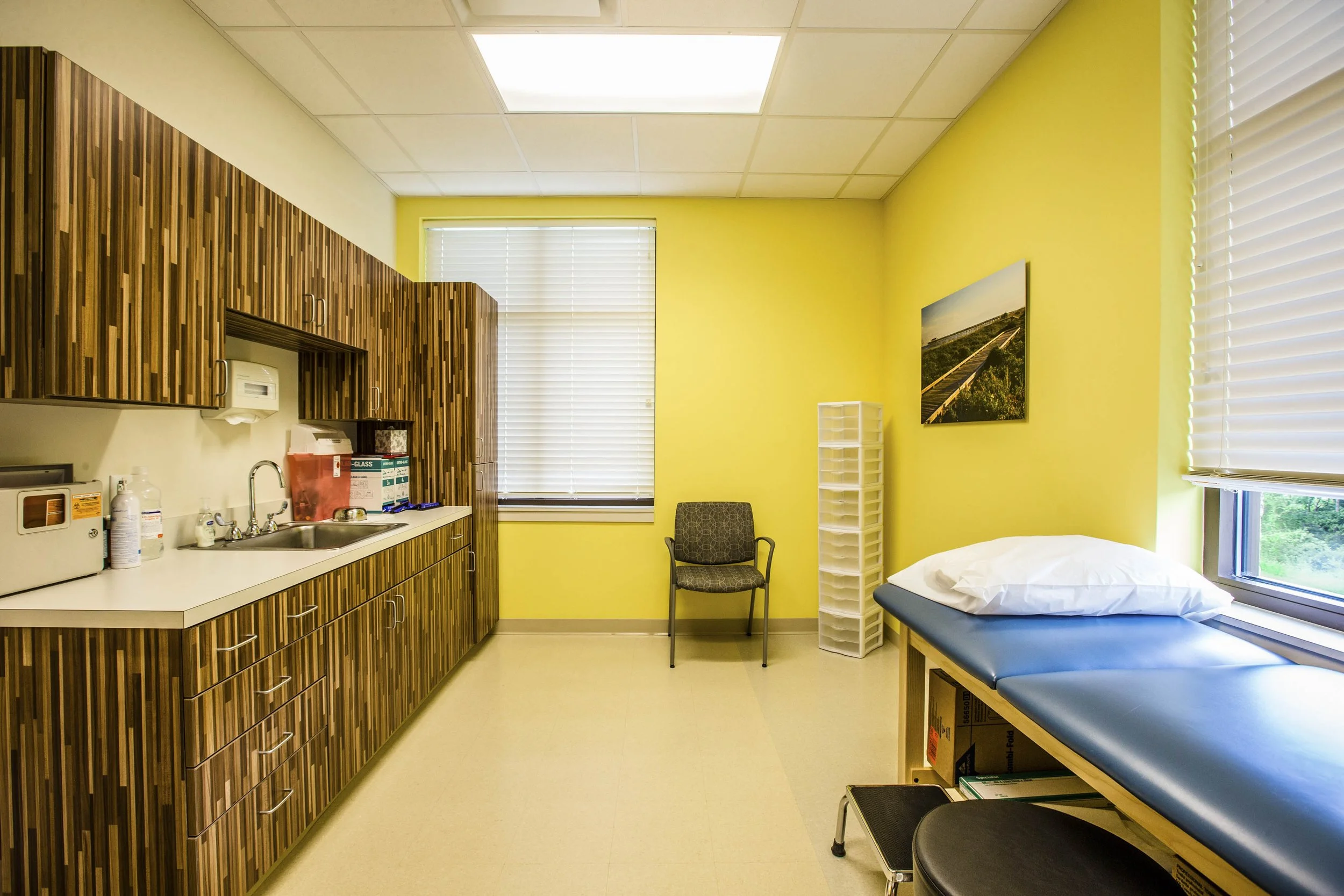 A medical examination room with yellow walls, a window with blinds, a countertop with medical supplies, a chair, a bed with white sheets, and a cabinet.