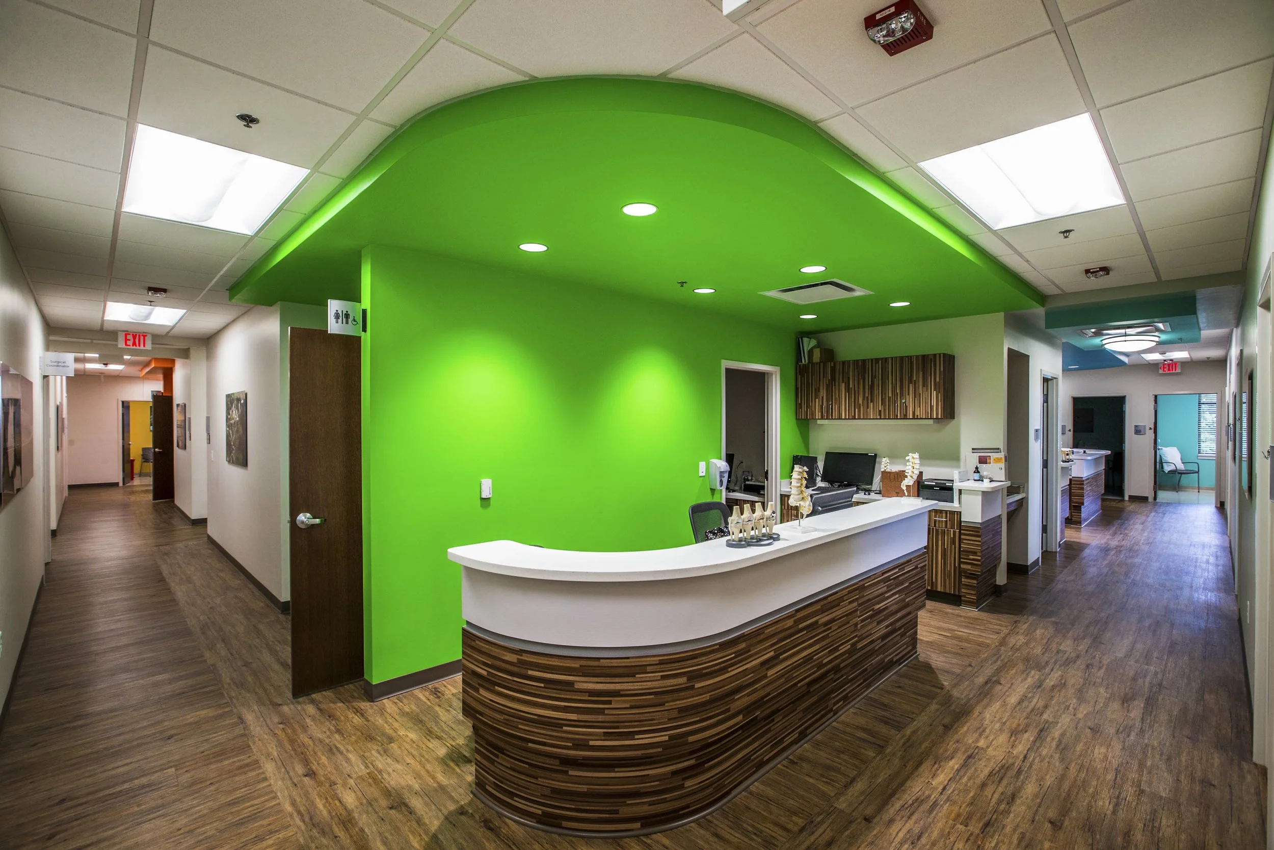 Hospital front desk with a curved wooden counter, green wall behind, and hospital staff area visible. Ceiling with bright lights, hallway with wooden flooring, and signs for restrooms and exits.