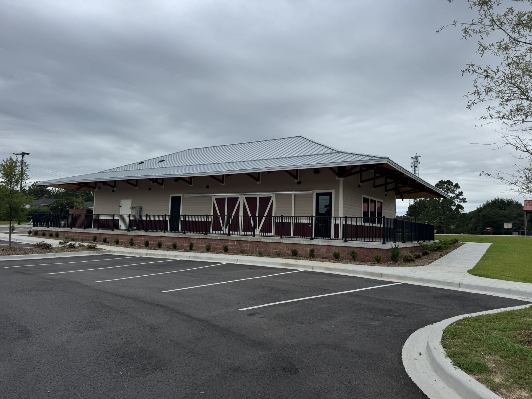 A small building with a metal roof and a porch, adjacent to an empty parking lot under a cloudy sky, with some trees and utility poles in the background.