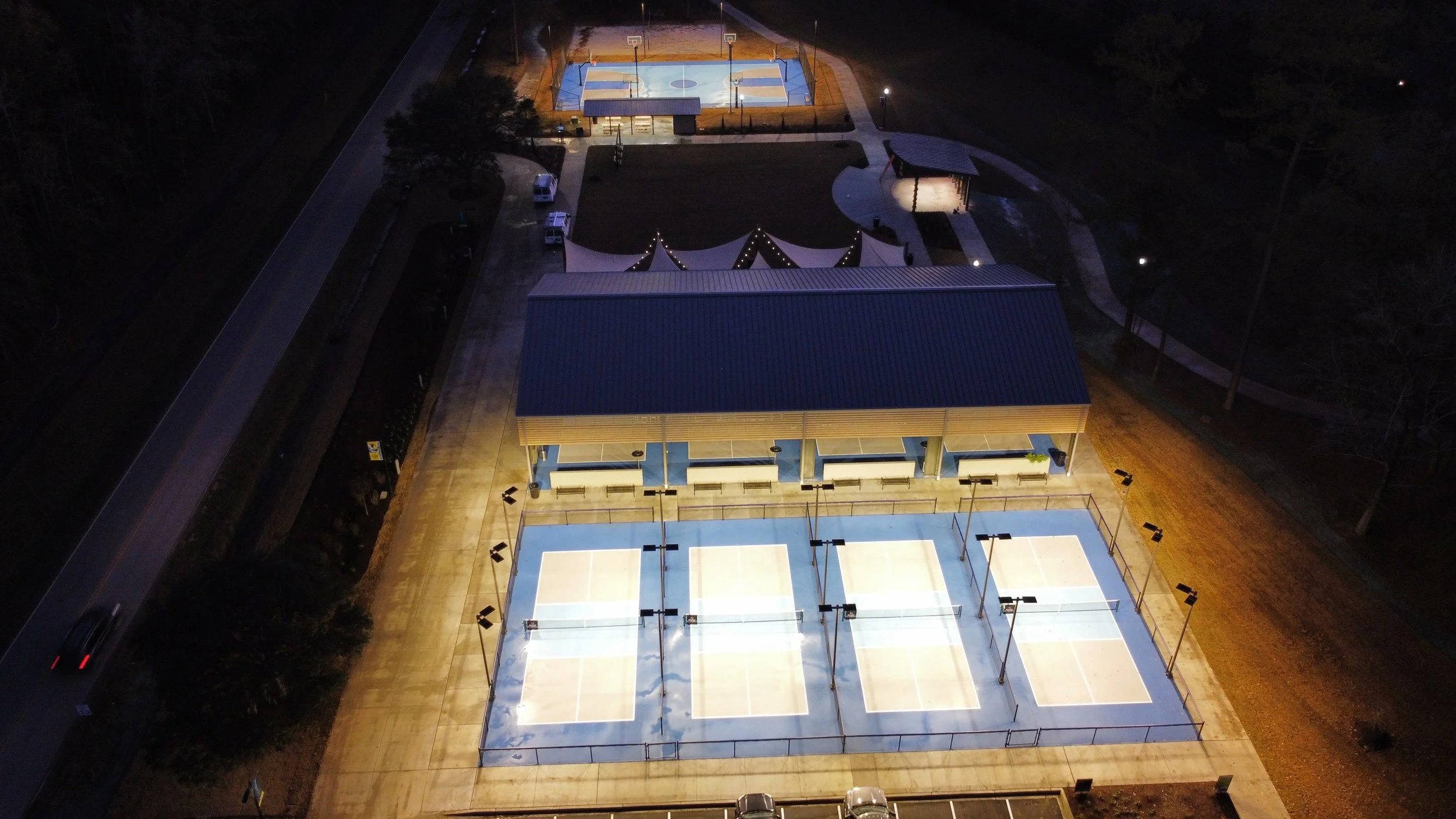 Aerial night view of a recreational park with tennis courts, basketball courts, a parking lot, and a playground illuminated by lights.