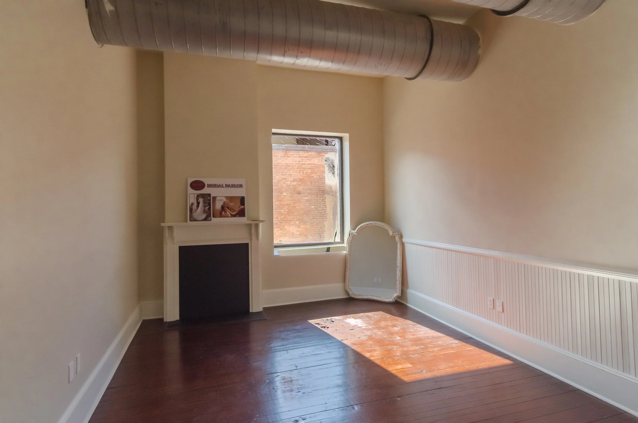 Empty room with hardwood floors, cream-colored walls, a fireplace with a white mantel, and a small window showing a brick building outside. There are posters on the wall and an ornate mirror leaning against the right wall.