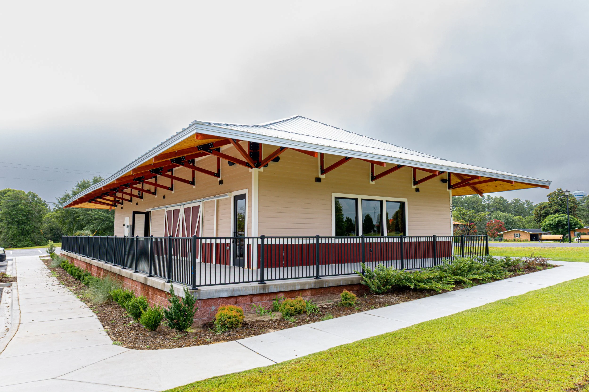 A modern building with a sloped metal roof, beige siding, and a black fence nearby. It features large windows and is surrounded by a landscaped yard with small bushes and grass. Overcast sky in the background.