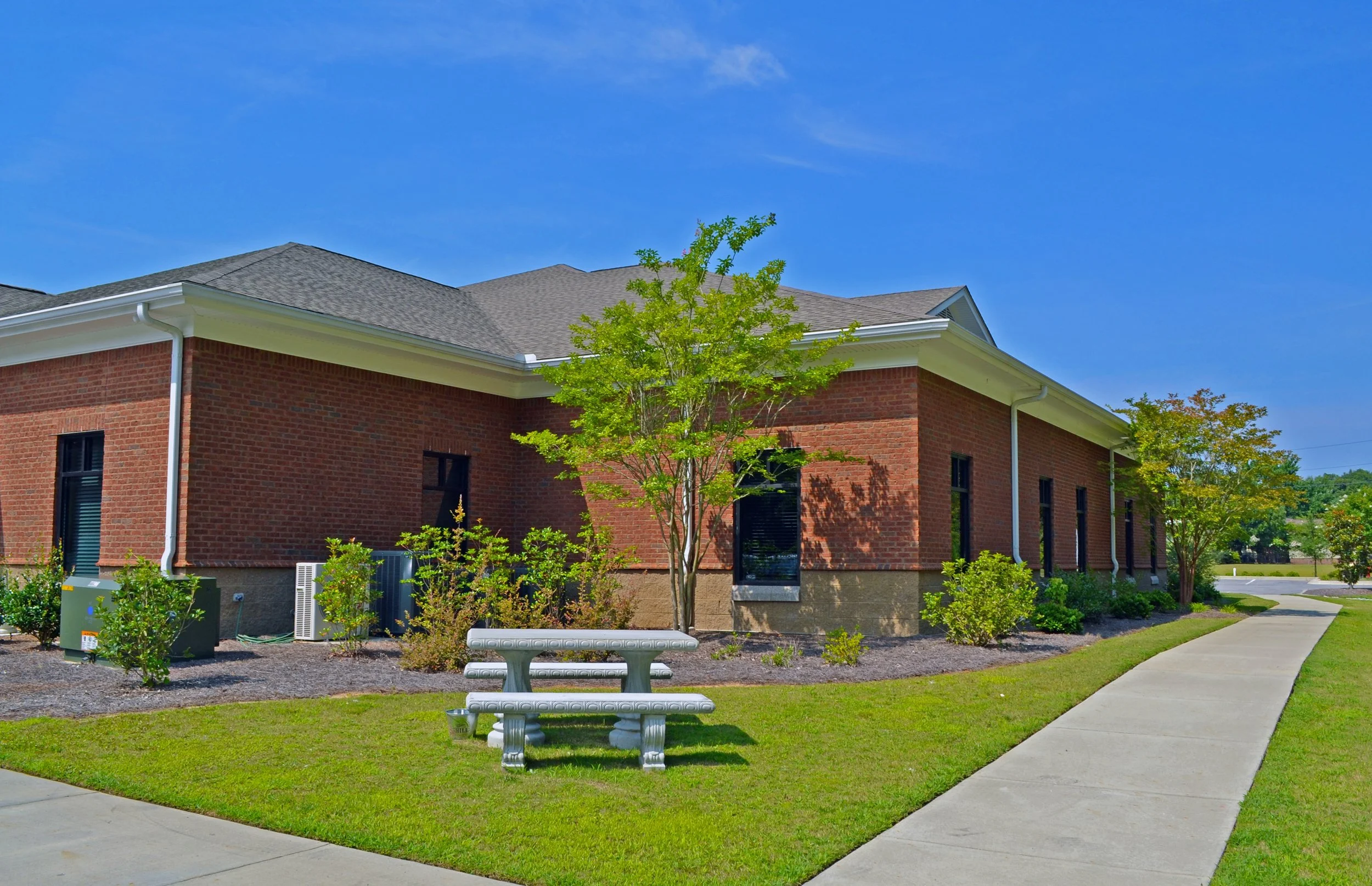 A brick building with a gabled roof and several windows, surrounded by a well-maintained lawn, trees, and a sidewalk with a bench.