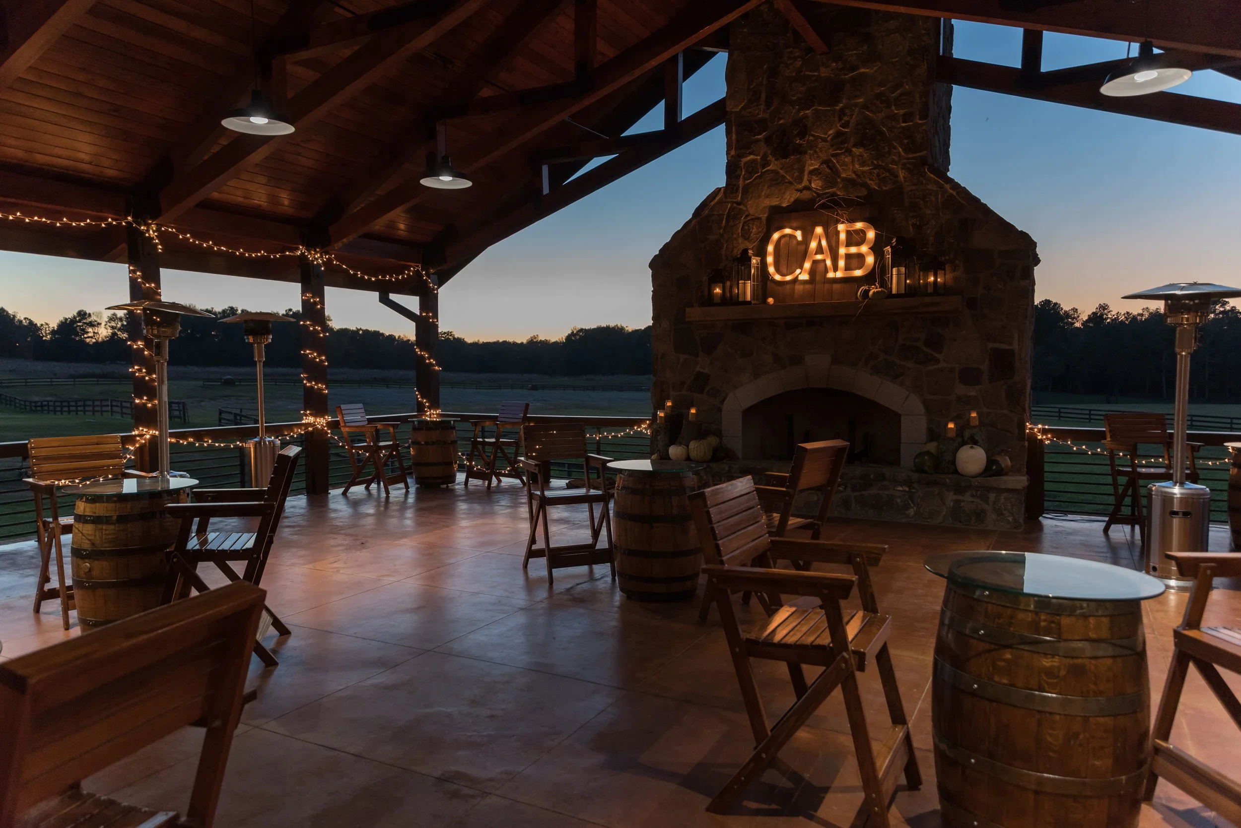 An outdoor patio area with wooden chairs, tables, lanterns, and a stone fireplace with a neon sign spelling 'CAB'. String lights and patio heaters are visible, and the background shows farmland and trees at dusk.