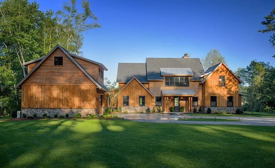 Large modern house with wooden exterior, surrounded by trees and a well-manicured lawn, under a bright blue sky.