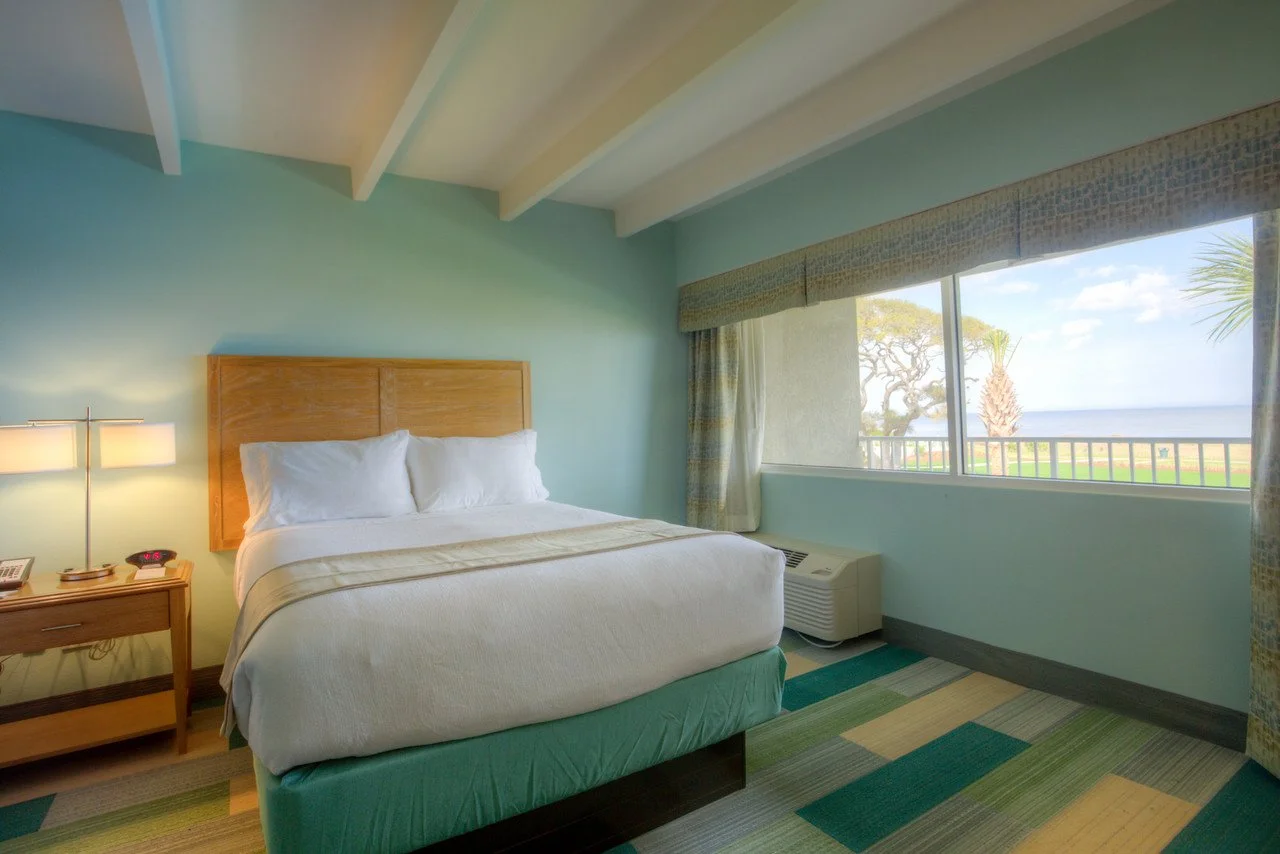 Hotel room with a bed, nightstand, lamp, and a large window showing a view of trees, grass, and the ocean.
