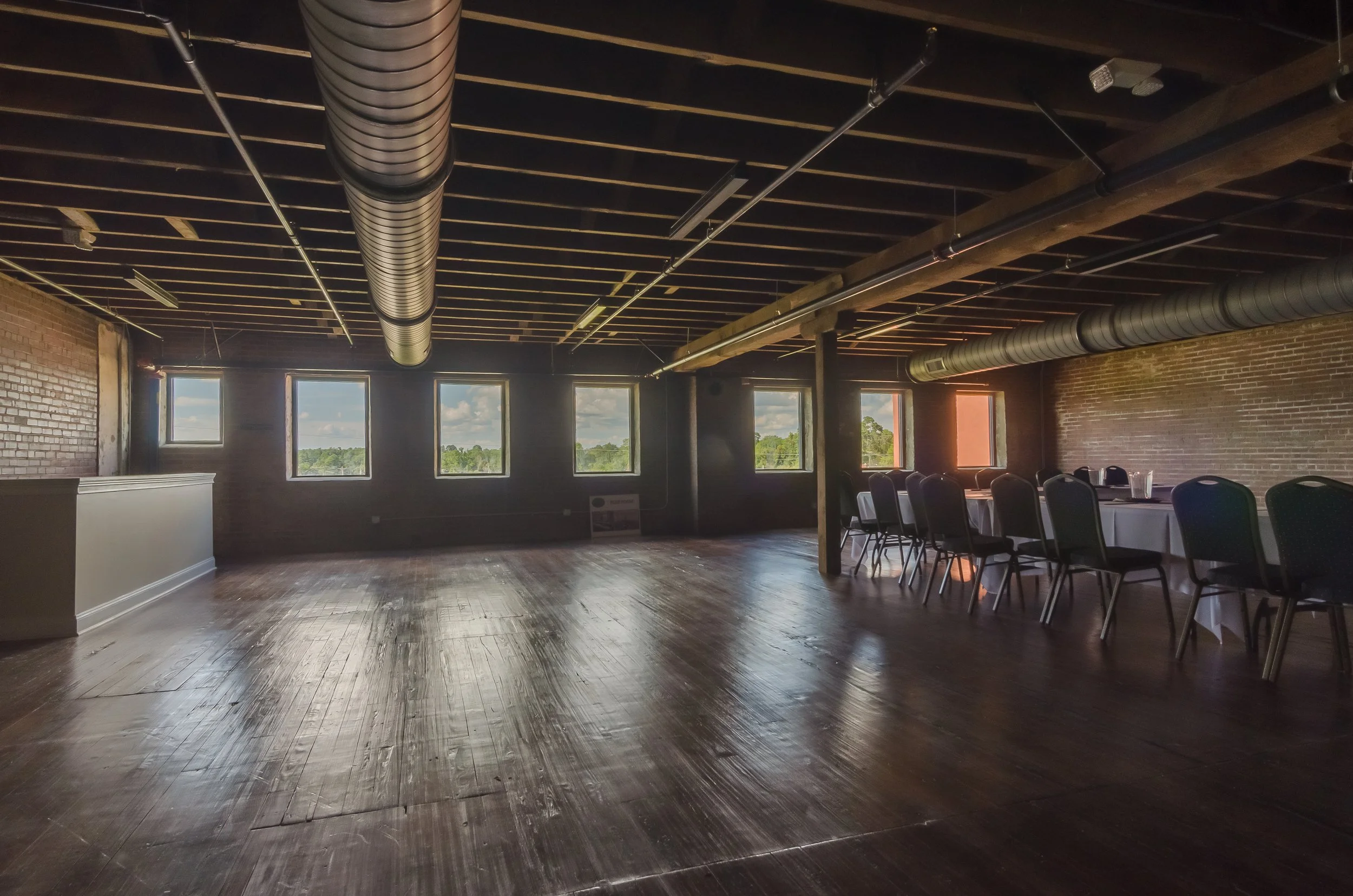 An empty event hall with dark wooden floors, brick walls, and large windows showing a scenic outdoor view. The ceiling has visible wooden beams, ductwork, and lighting fixtures. A long table with chairs is set up to the right.