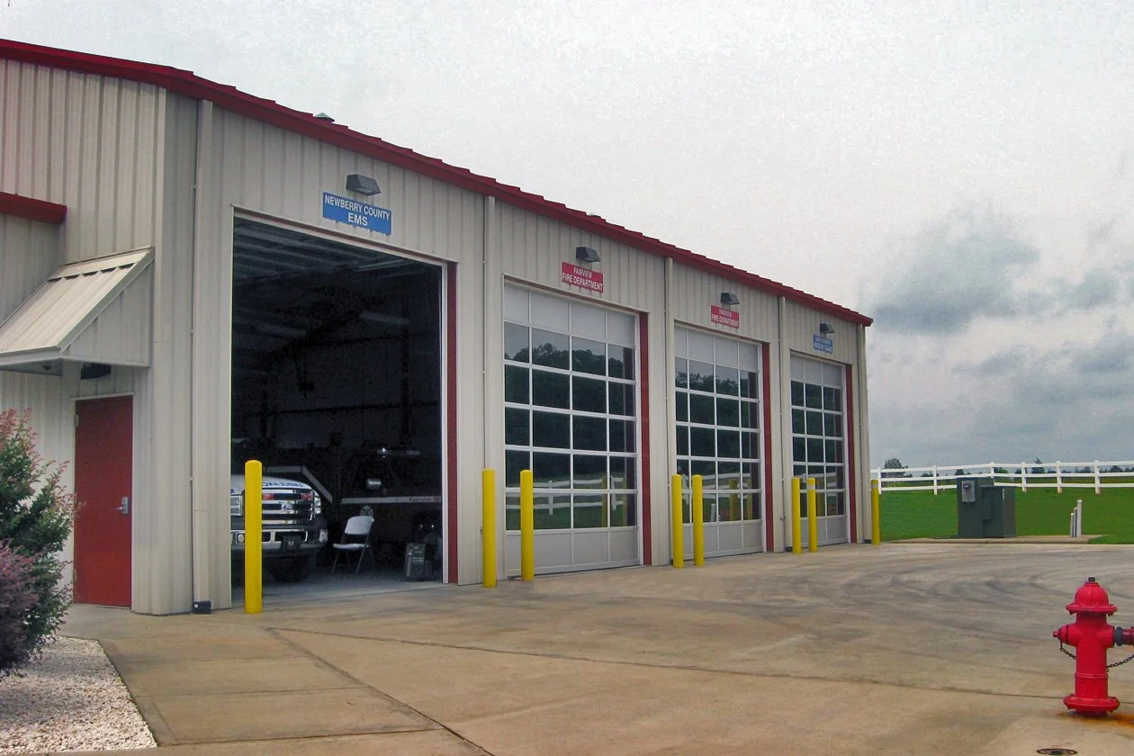 Fire station with four large garage doors, one open revealing an ambulance inside, blue and red signage above each door, yellow safety posts, an adjacent red door, and a small landscape with a bush and a fire hydrant.