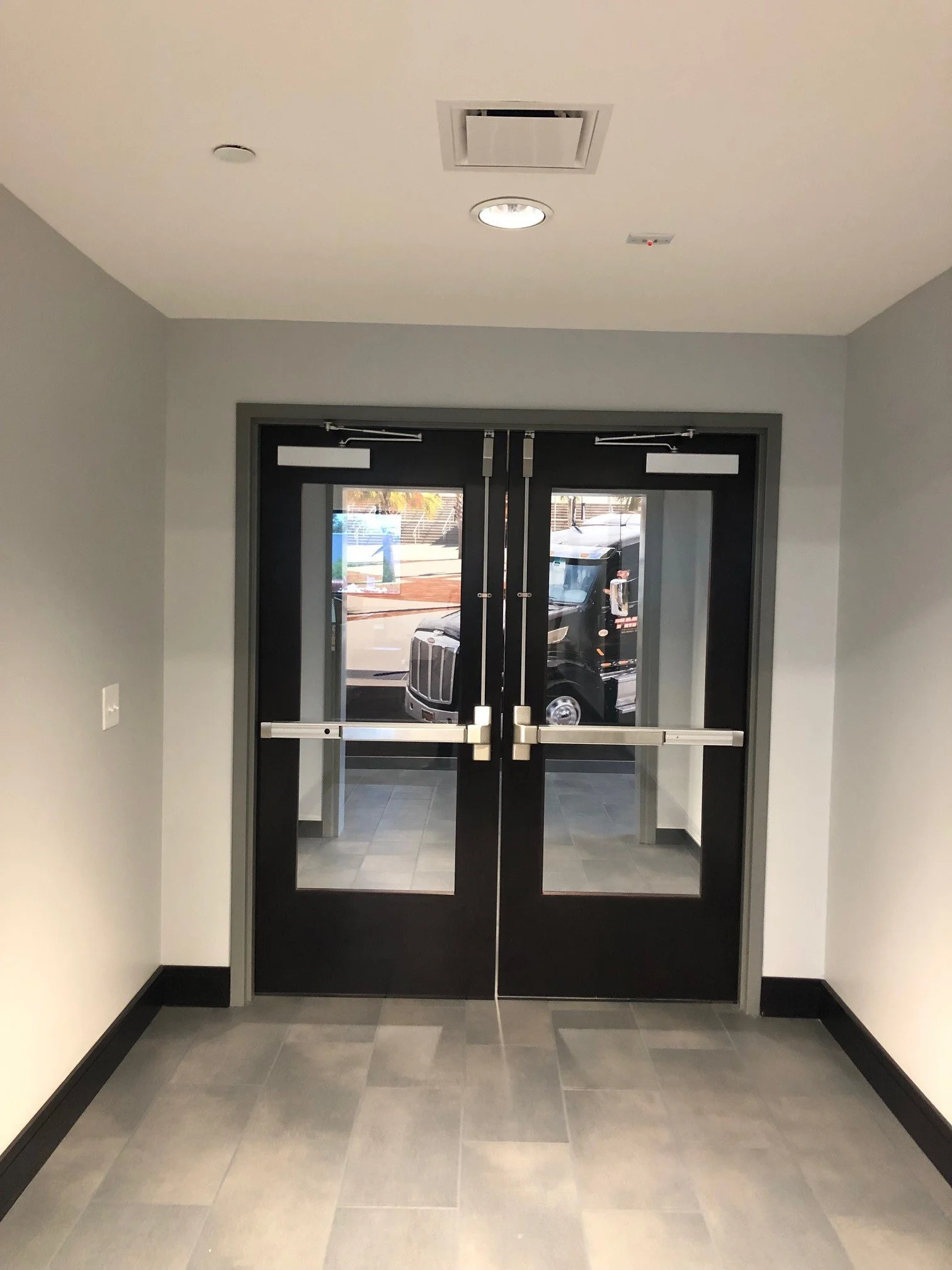 Interior view of a building with double glass doors leading outside, visible cars, and sidewalk scene through the windows. The door is black with metal hardware, and the walls are painted light gray with black trim.