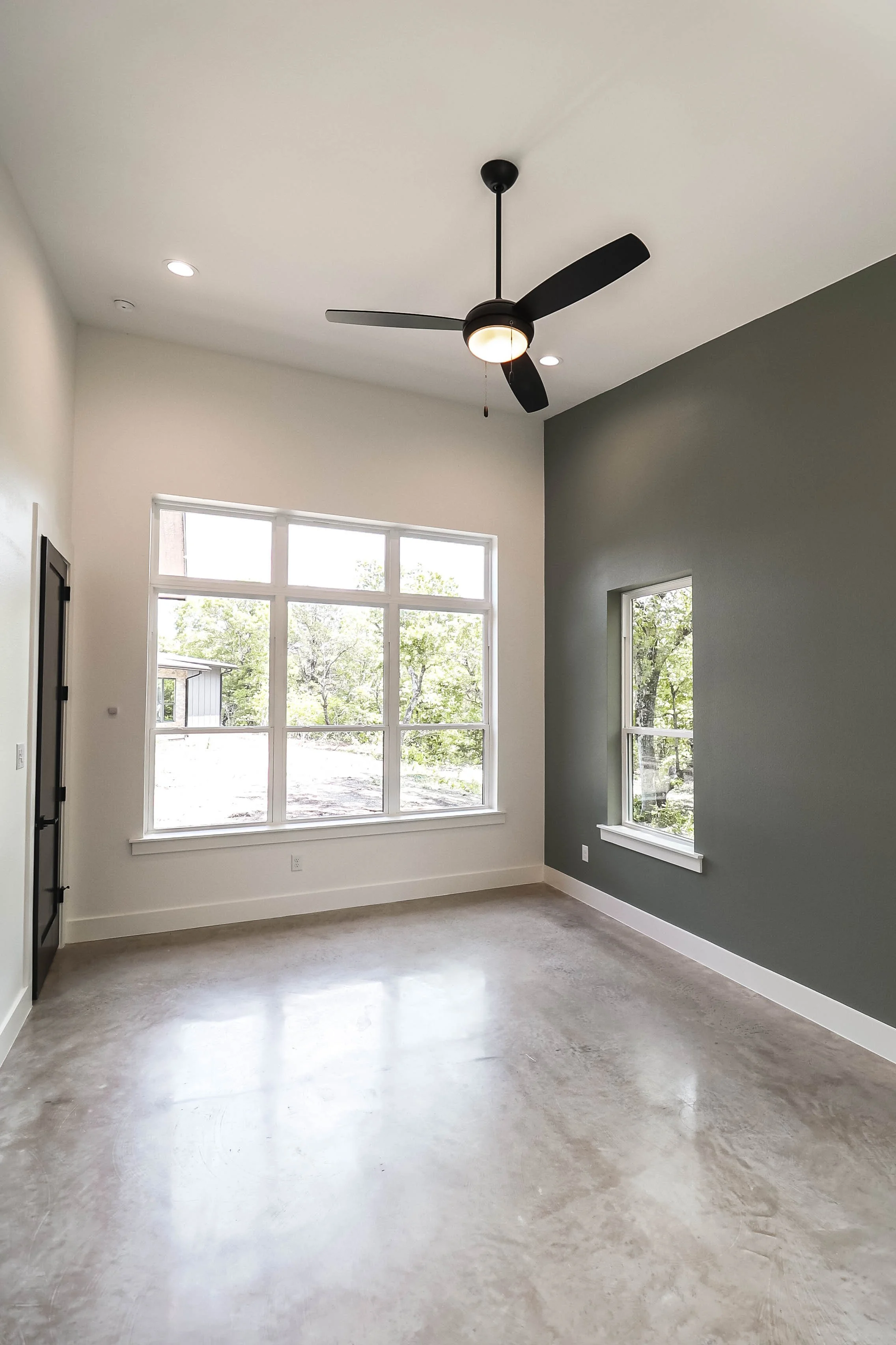 Empty room with large windows, a ceiling fan with lights, and a gray accent wall, with natural light coming in.