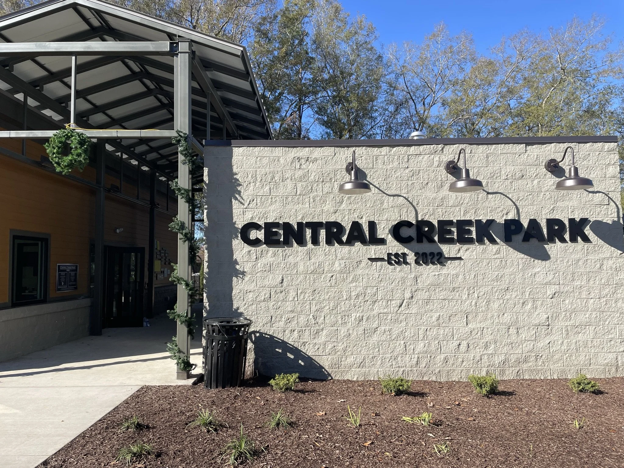 Entrance of Central Creek Park with a white brick wall, black sign, decorative lights, a trash can, some small plants, and a sidewalk leading inward. Part of the building with greenery decorations and an overhang is visible on the left.