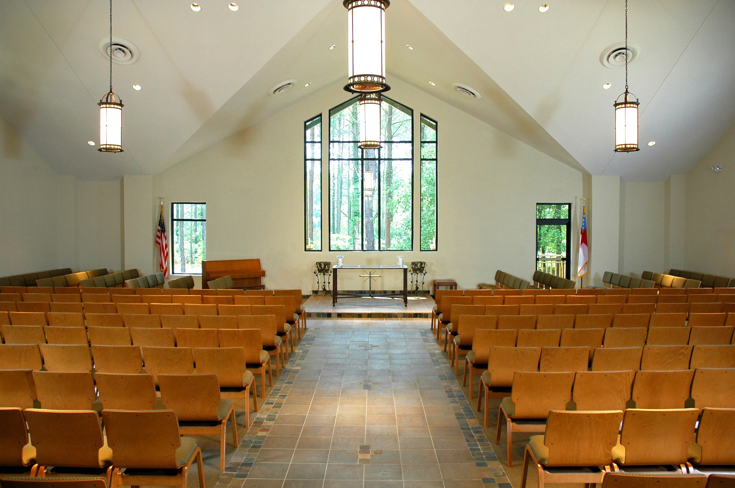 Interior of a church or chapel with rows of wooden chairs, a raised altar with a cross, flags on either side, large stained glass windows, and hanging pendant lights.