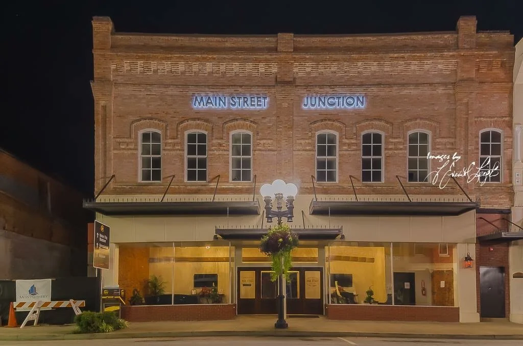 A historic brick building at night with neon signs reading 'Main Street' and 'Junction'. The ground floor has large windows with warm interior lighting, and a streetlamp with hanging plants is in front of the building.