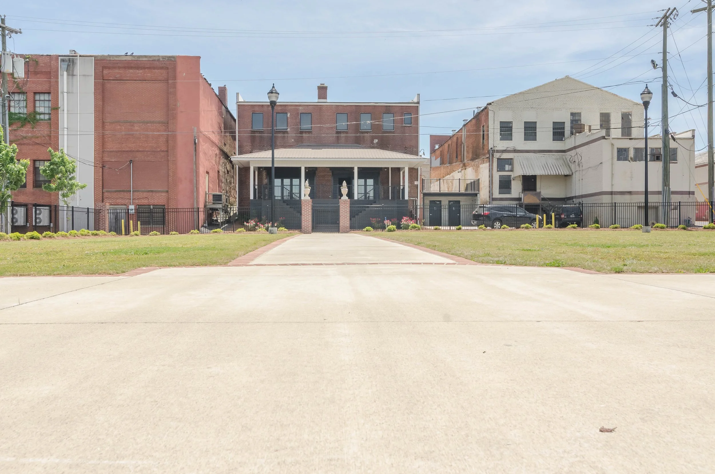 An empty paved area with a grassy border leading up to a fenced park with buildings in the background.