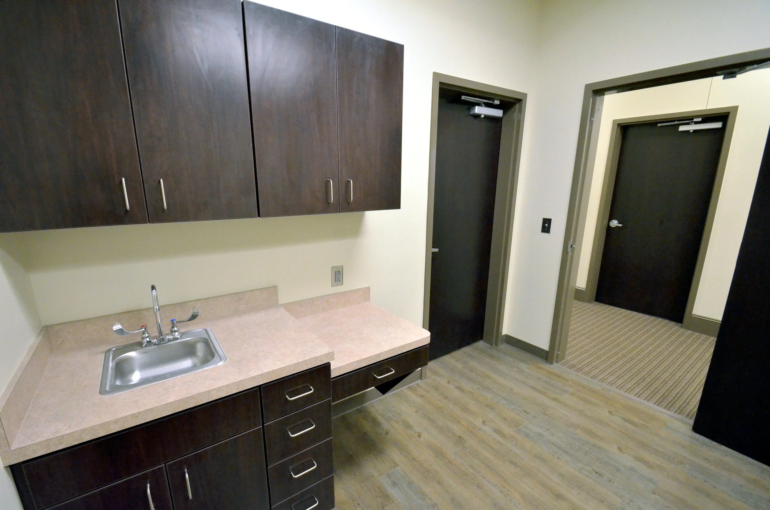 Image of a kitchen area with dark wood cabinets, a beige countertop, a stainless steel sink, and two doors leading to separate rooms. The flooring is wood laminate.