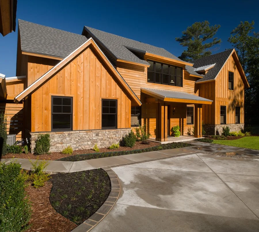 Modern two-story house with wooden exterior, stone accents, and a covered front porch, surrounded by a well-maintained landscape.