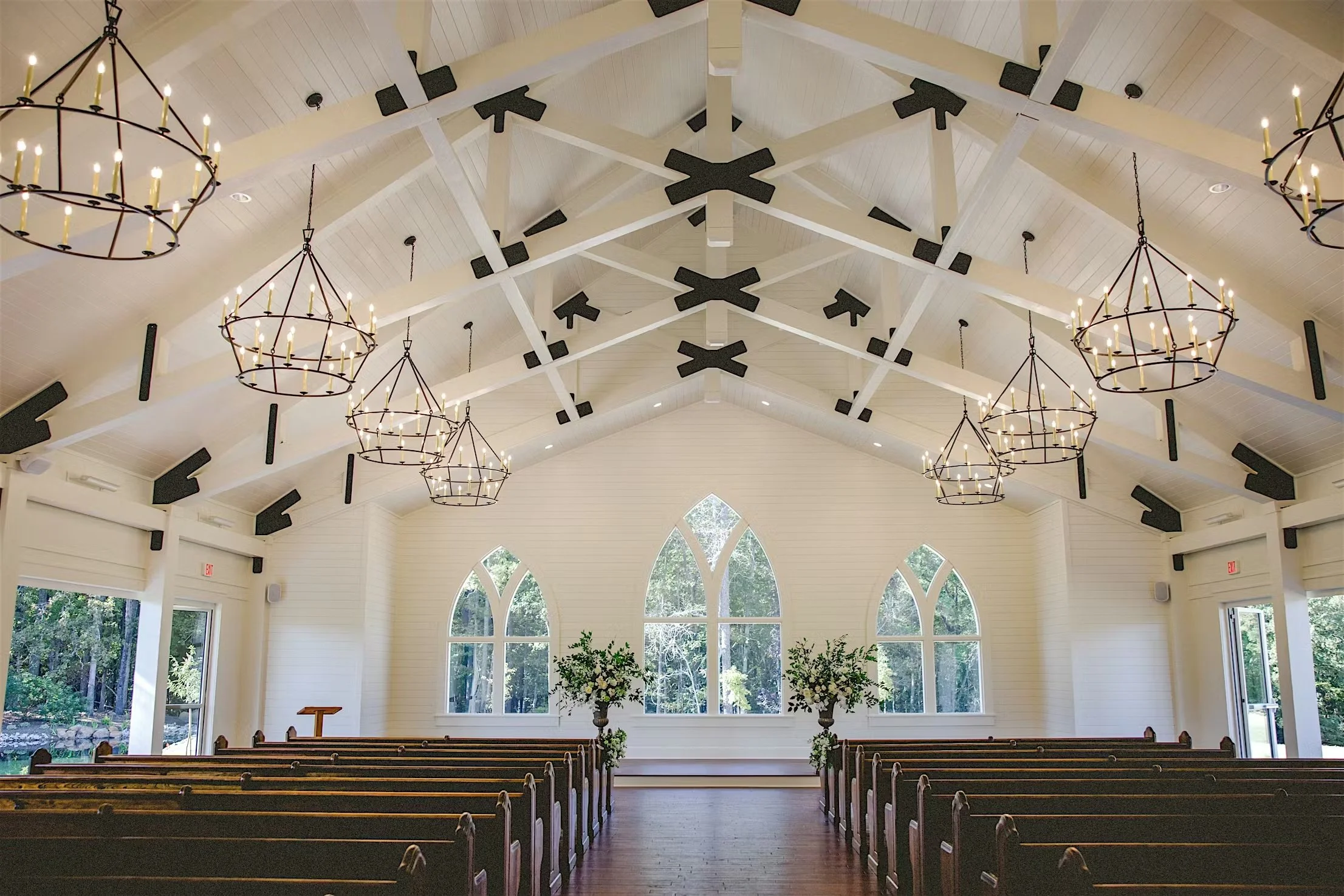 Interior of a church with white wooden walls and high ceiling, dark wood pews, large pointed arch windows, and black and gold chandeliers hanging from the ceiling.