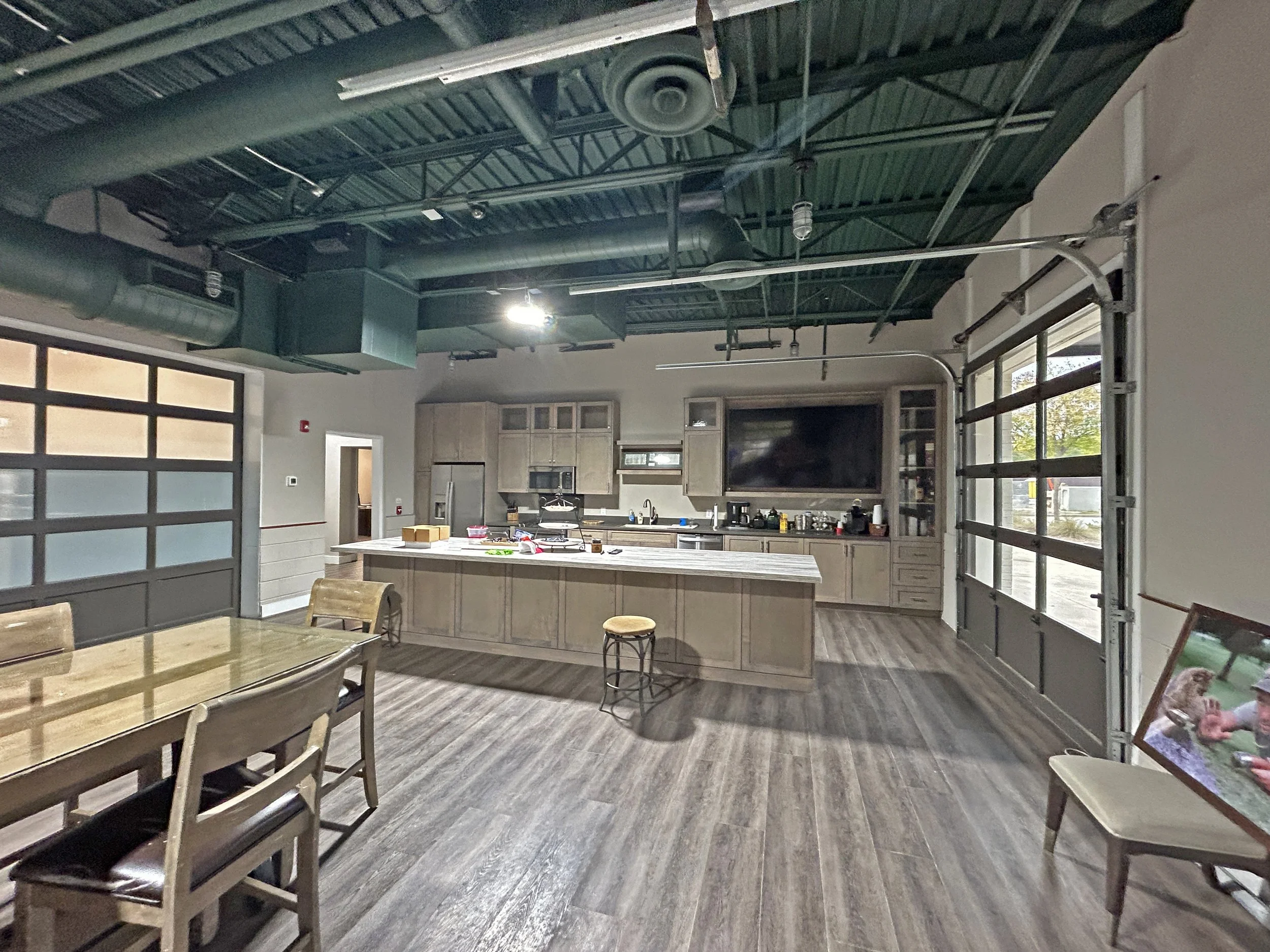 A modern, industrial-style kitchen with a high ceiling featuring exposed green metal ductwork, an island with a light wooden top, and several chairs around it. There are large windows bringing in natural light, and stainless steel appliances in the b