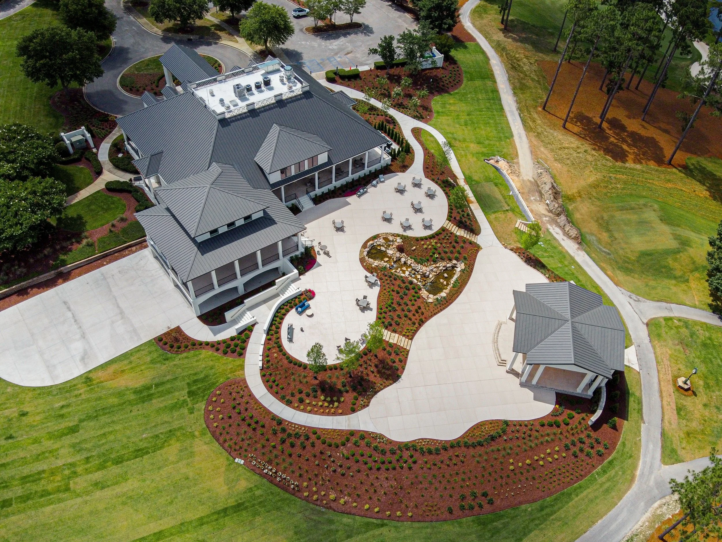 A large building with a gray metal roof, surrounded by landscaped gardens with flower beds and trees, and a paved patio with outdoor seating. There are paved walkways, a parking lot, and green grassy areas.