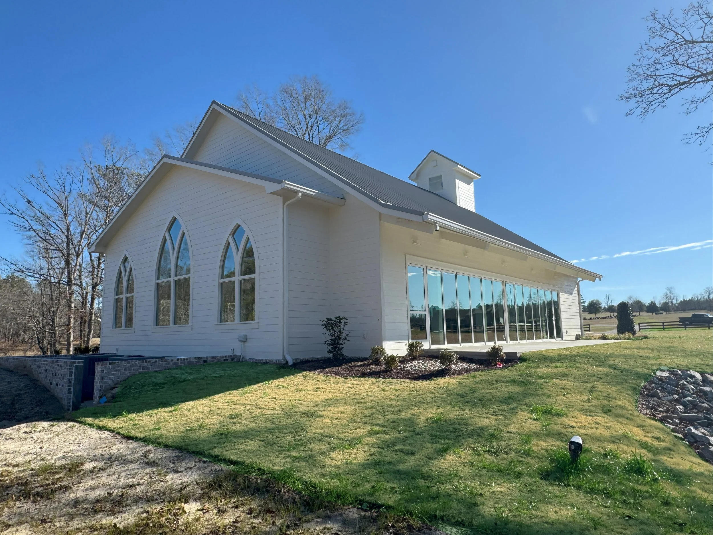 A white church with arched windows and a tall roof, surrounded by trees and a grassy yard, under a clear blue sky.