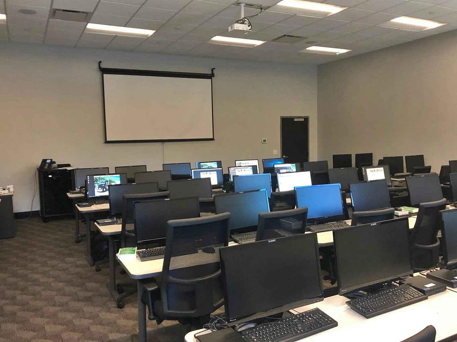 Empty classroom or training room with multiple desktop computers on long tables, black chairs, a large projection screen at the front, and a ceiling-mounted projector.