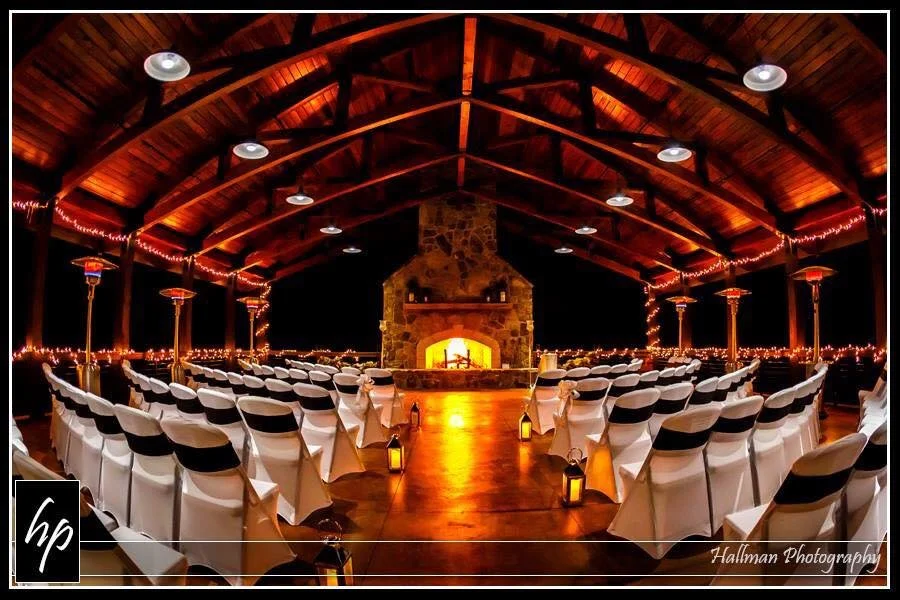 Indoor event space with rows of white chairs facing a stone fireplace with a fire, wooden ceiling with string lights, and small lanterns on the floor.