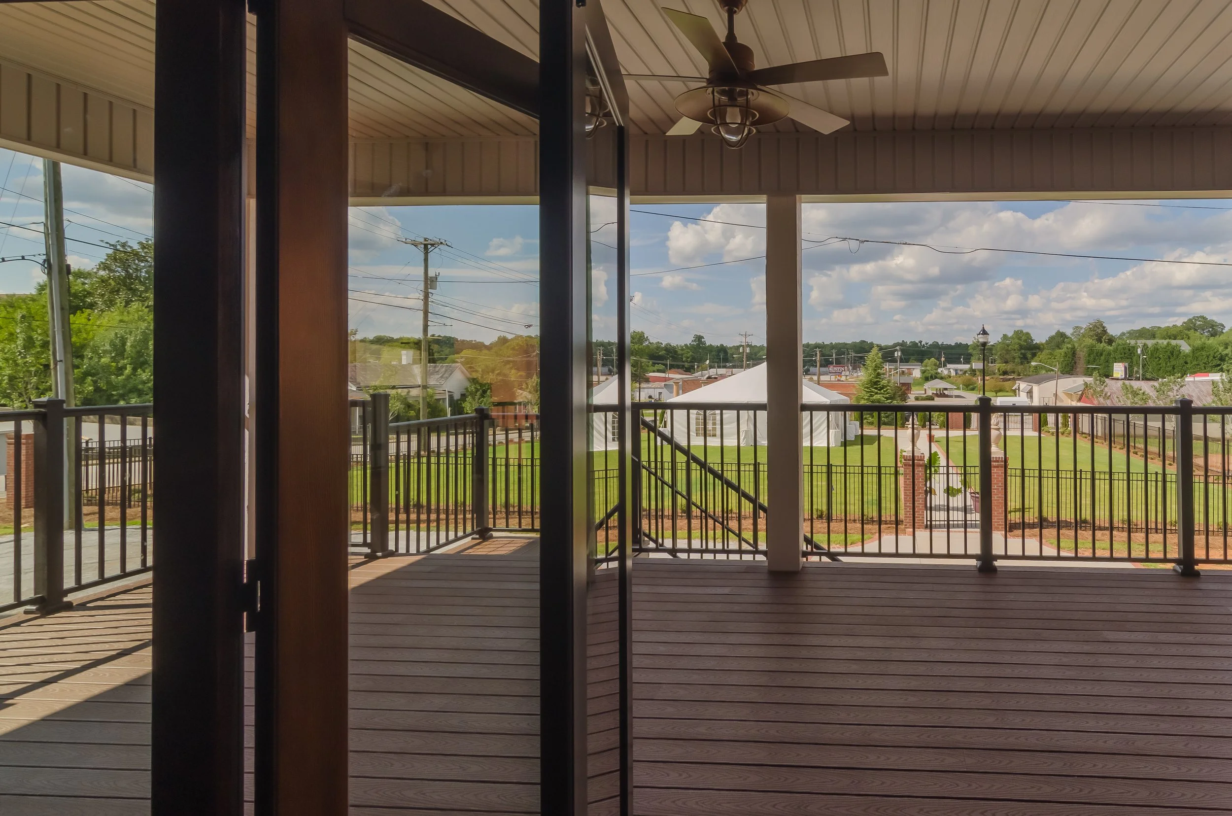 View from a covered balcony showing a backyard with green grass, a white canopy tent, a black metal fence, and surrounding houses under a partly cloudy sky.