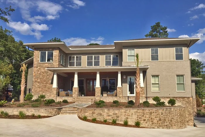 Modern two-story house with stone and siding exterior, multiple windows, a front porch with columns, and a landscaped front yard with palm trees.