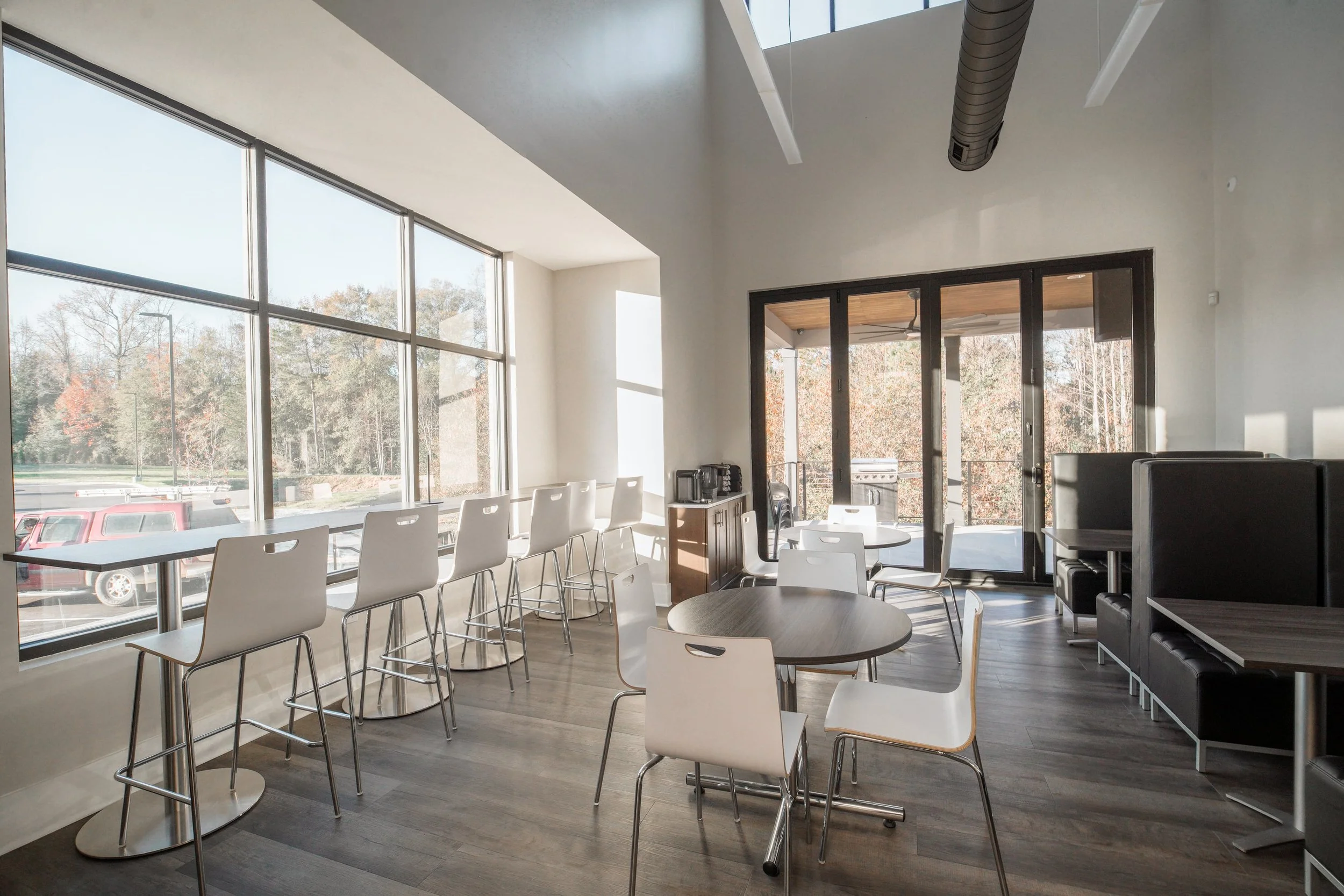 Bright dining area with large windows, white chairs, round tables, and a view of trees outside.