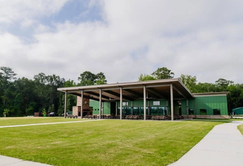 A modern green building with a large outdoor covered patio, surrounded by a grassy area and trees in the background.