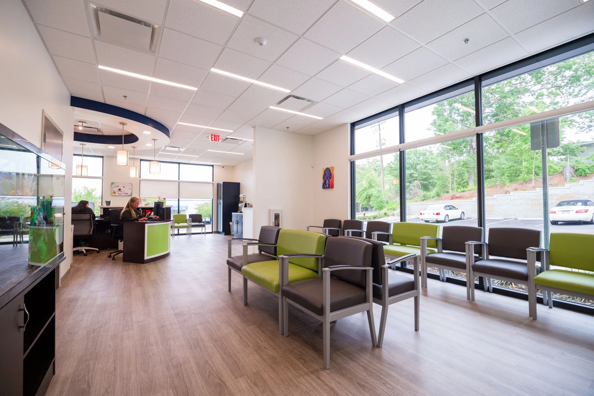 A brightly lit waiting area with large windows showing green trees outside, multiple chairs with green and gray cushions, a reception desk with two employees working, and artwork on the walls.