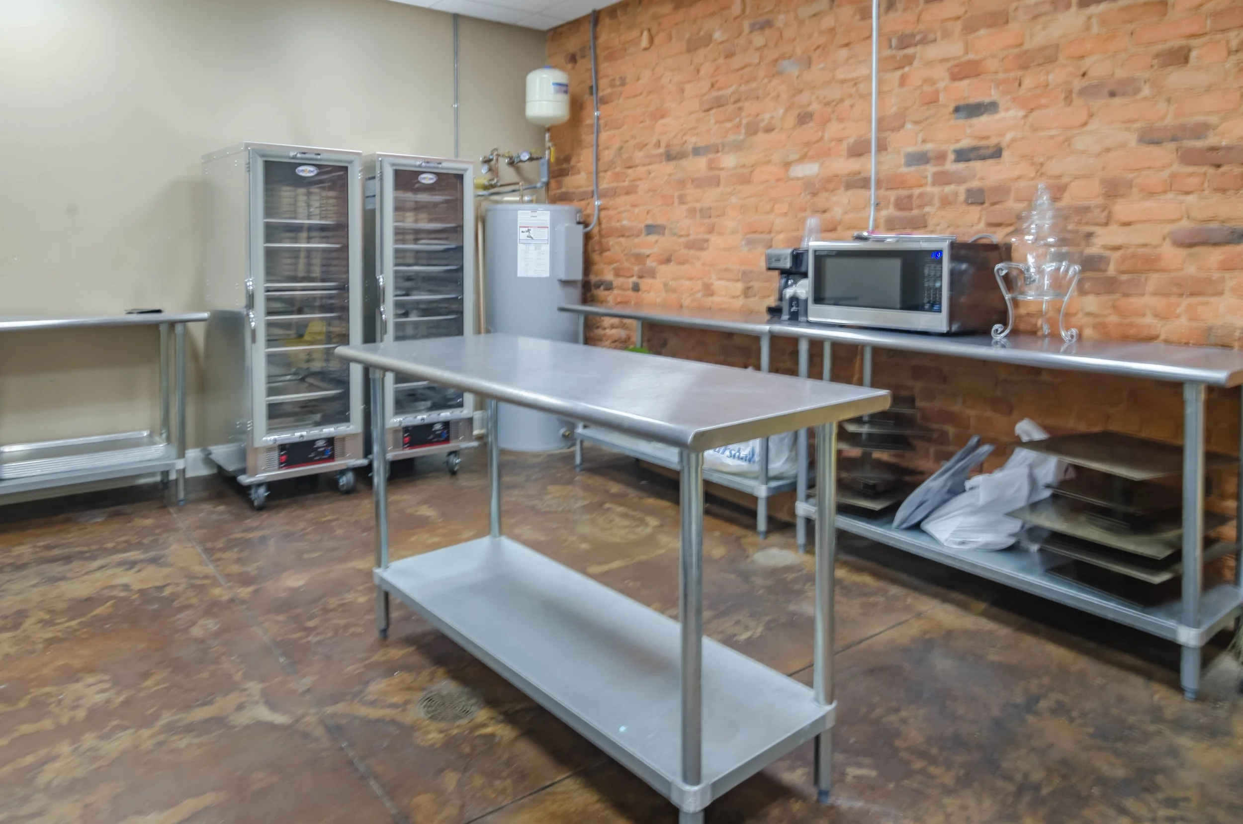 Commercial kitchen or bakery equipment storage area with stainless steel tables, two refrigerated display cases, a microwave, and a small glass bowl on a brick wall background.